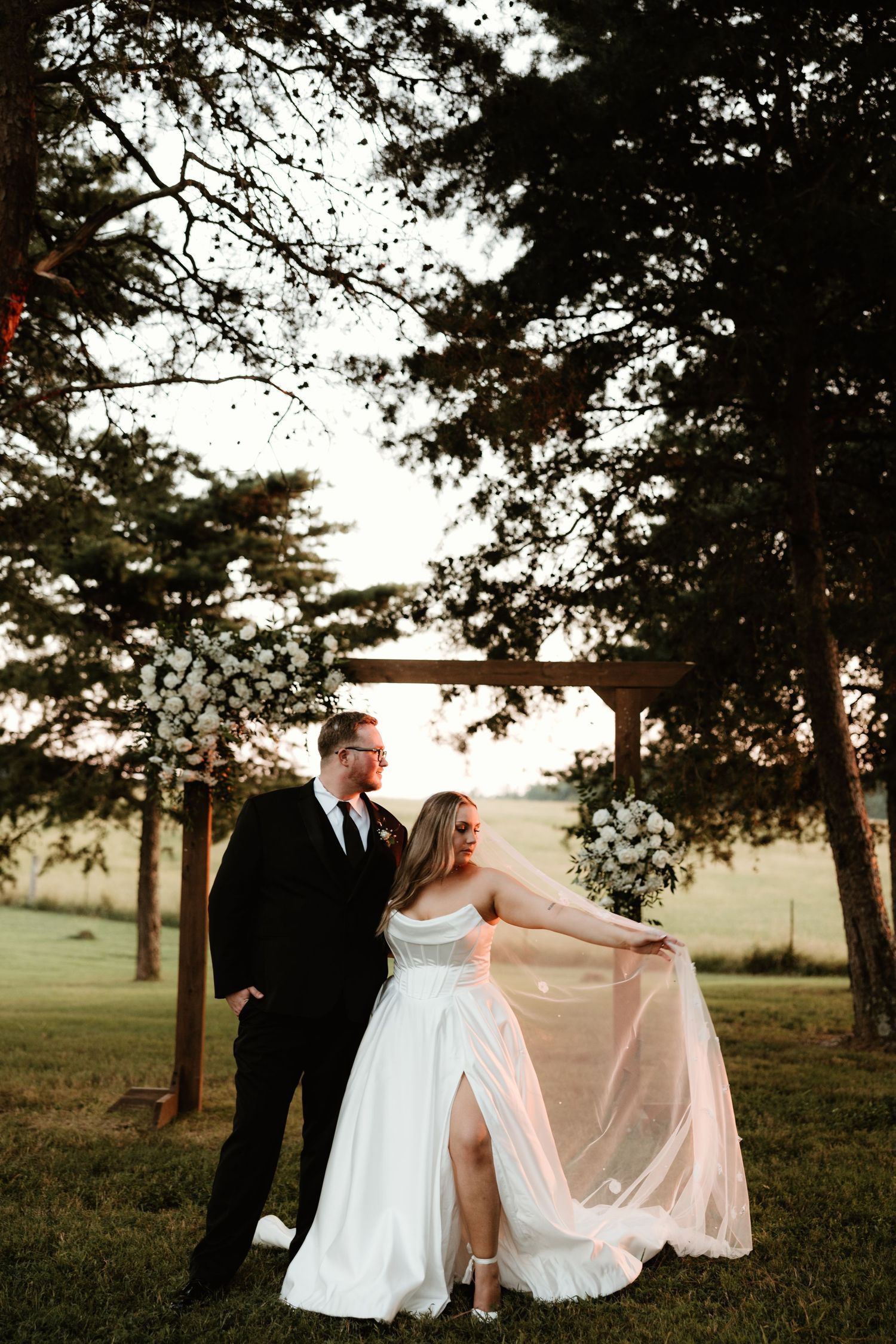 A romantic outdoor wedding photo with couple standing beneath trees at sunset while bride holds flowing veil.
