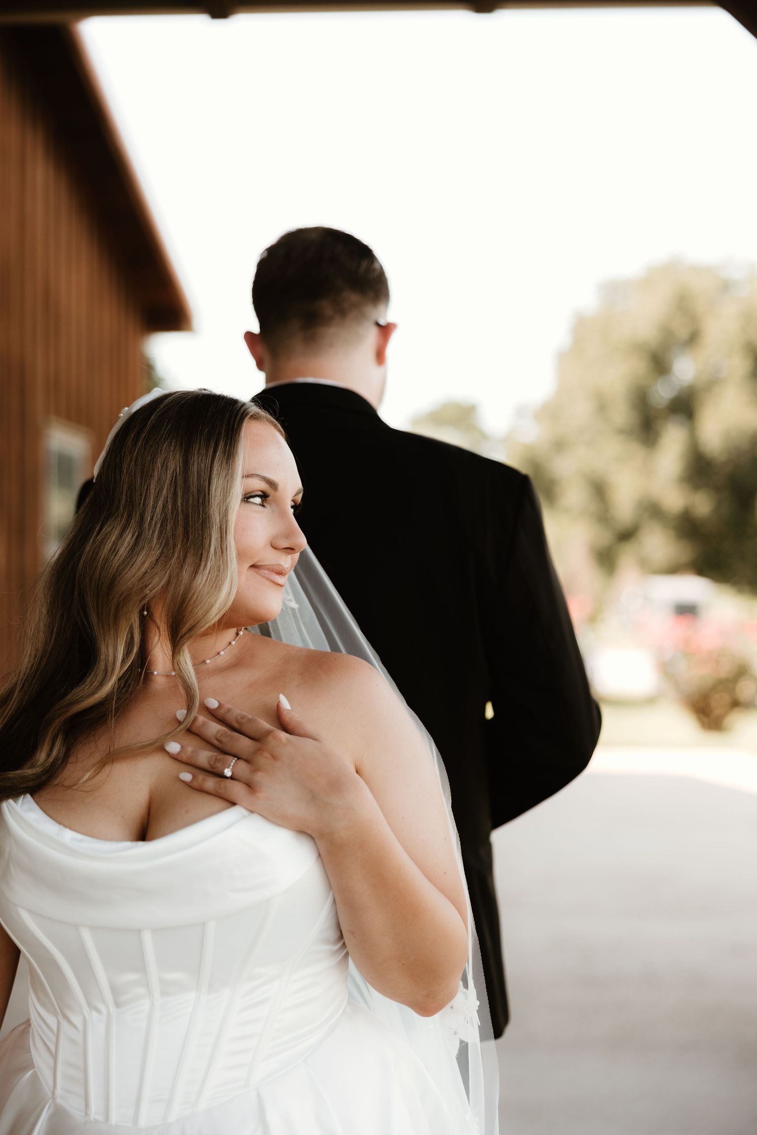 A bride in a white wedding dress smiles over her shoulder while showing her ring as her groom faces away.