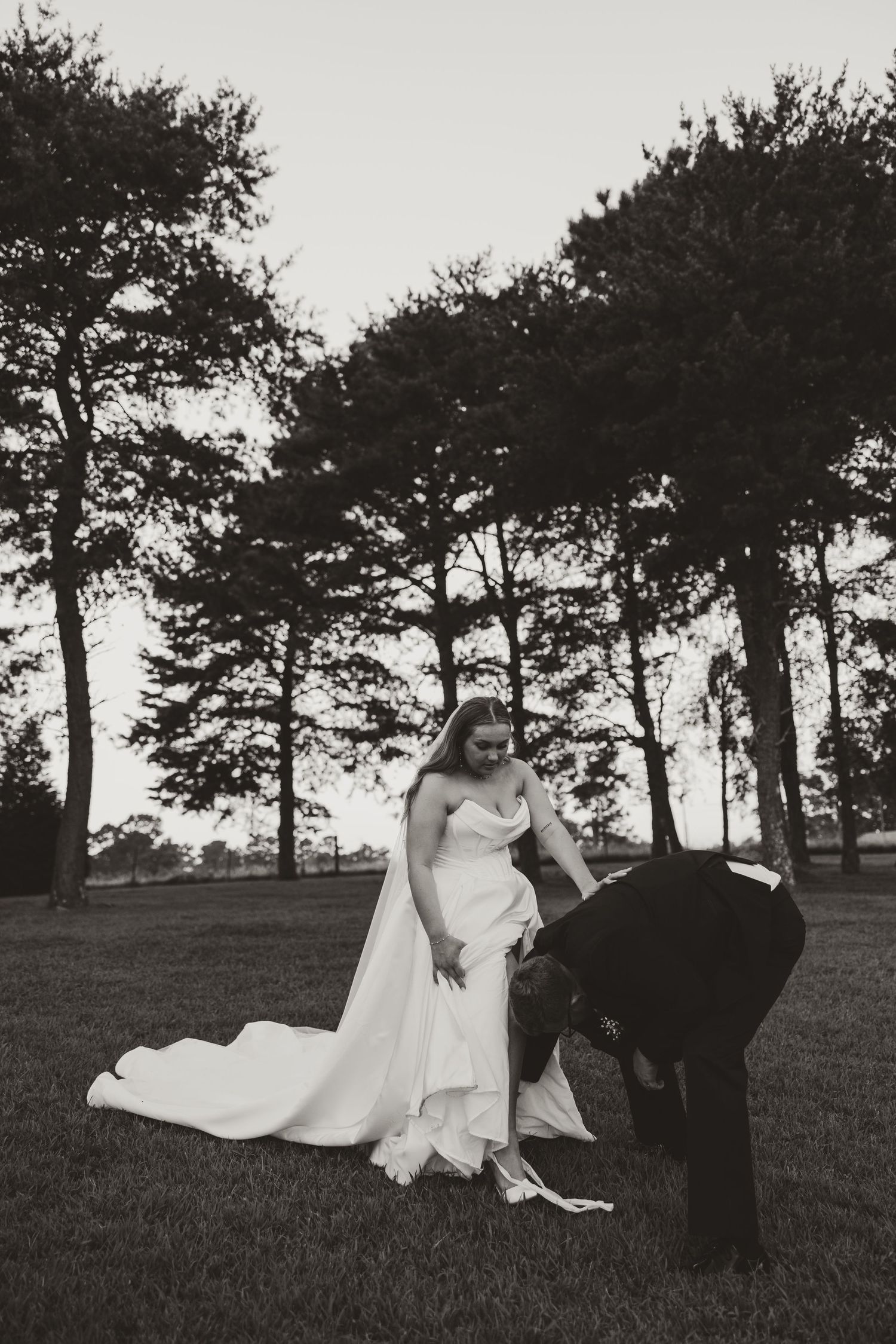 Romantic black and white photo series showing a couple embracing on a grassy hill with tall pine trees in the background.