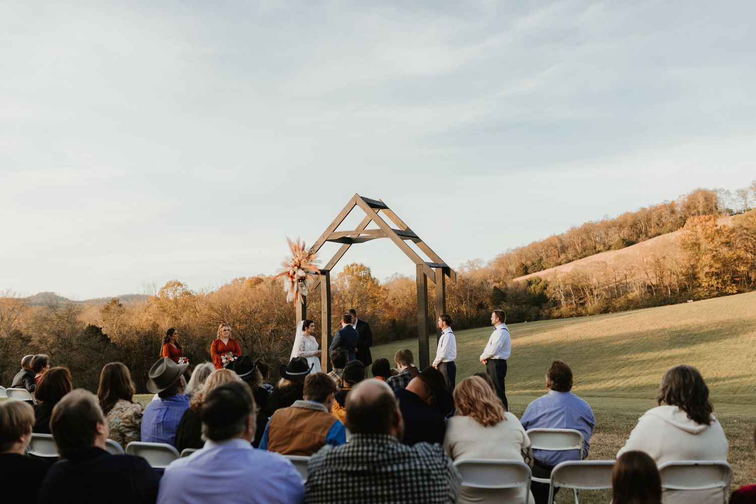 A line of wedding guests form archways with their arms raised outdoors against a sunset sky.