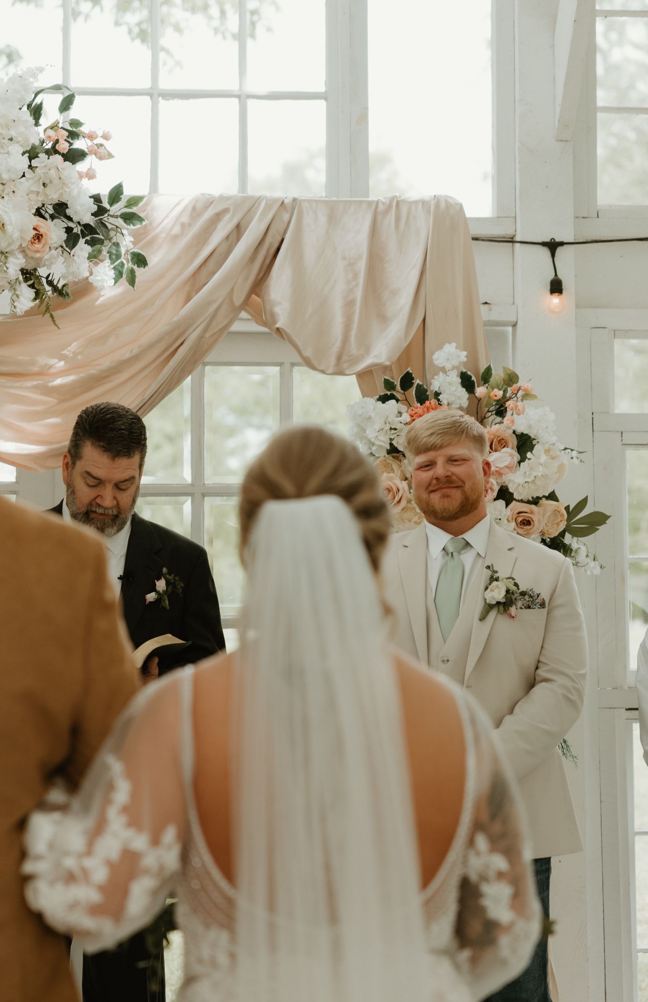 Wedding ceremony in a light-filled room with white drapery and floral arrangements creating an elegant archway.