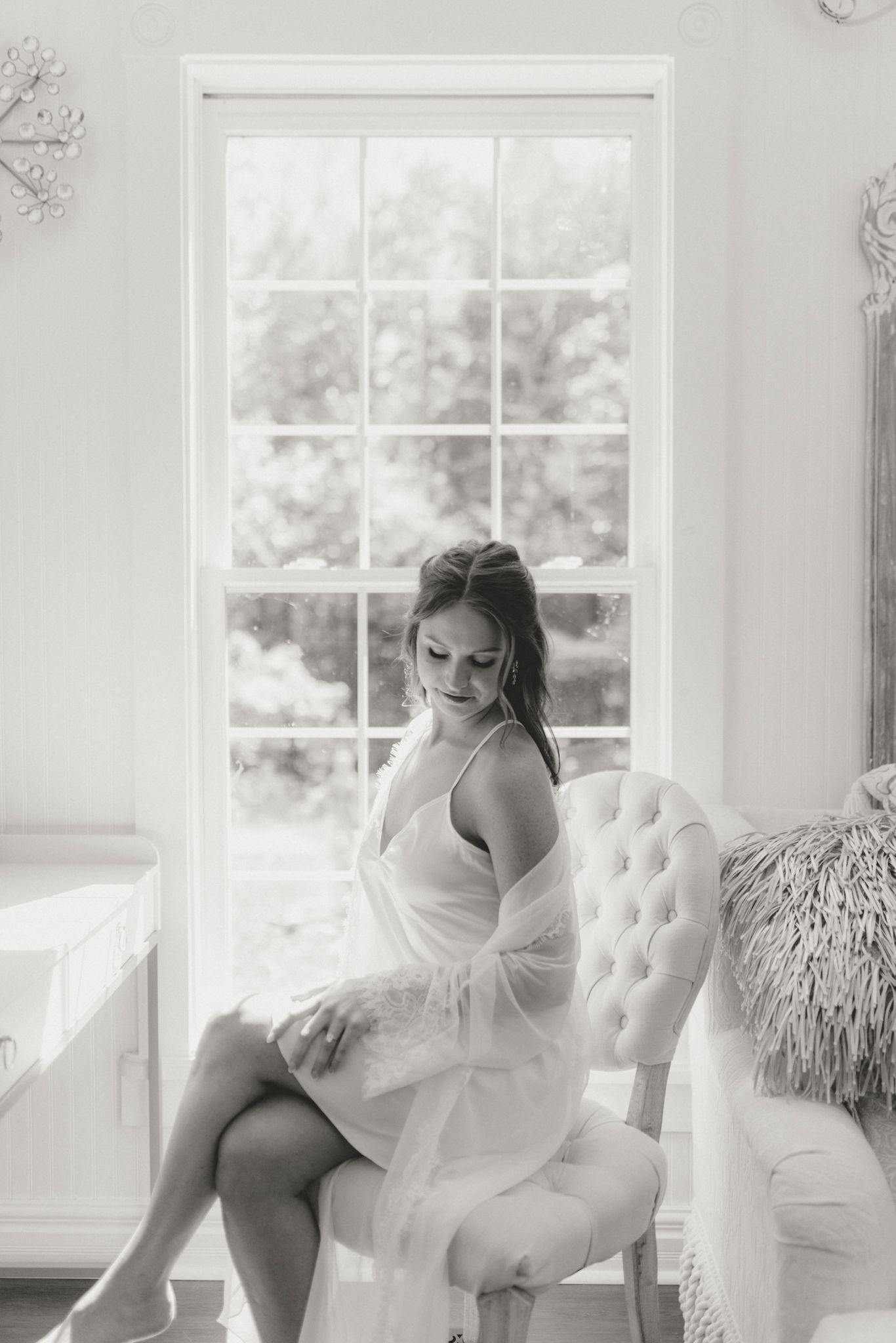 Soft boudoir photo near bright window with figure seated in tufted white chair.
