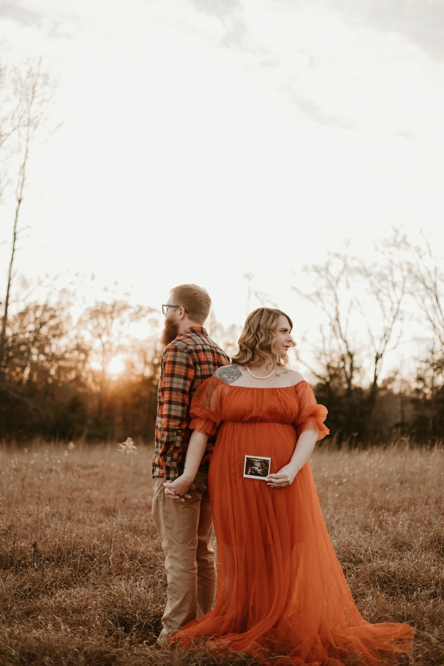 Couple in autumn field holding sonogram photo and embracing at sunset in warm orange and brown tones.