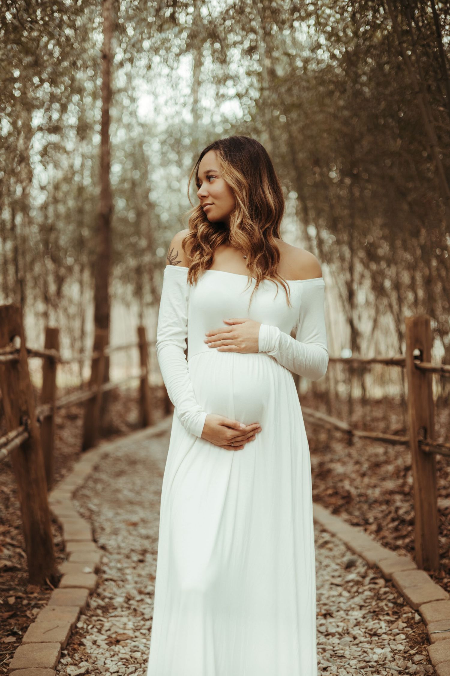 A maternity photoshoot in the woods features an off-shoulder white dress against a dreamy forest backdrop.
