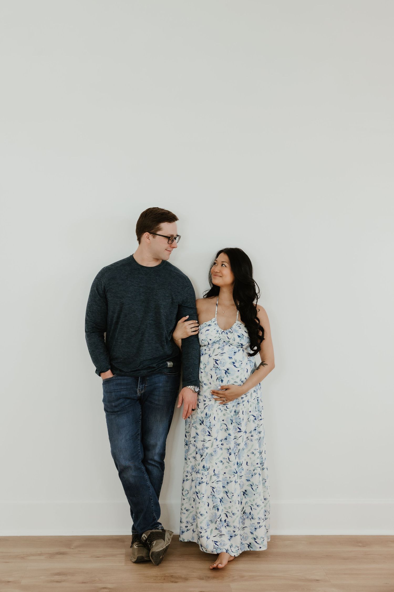 Couple posing against white wall, pregnant woman in floral dress cradling baby bump, man in dark sweater and jeans smiling.