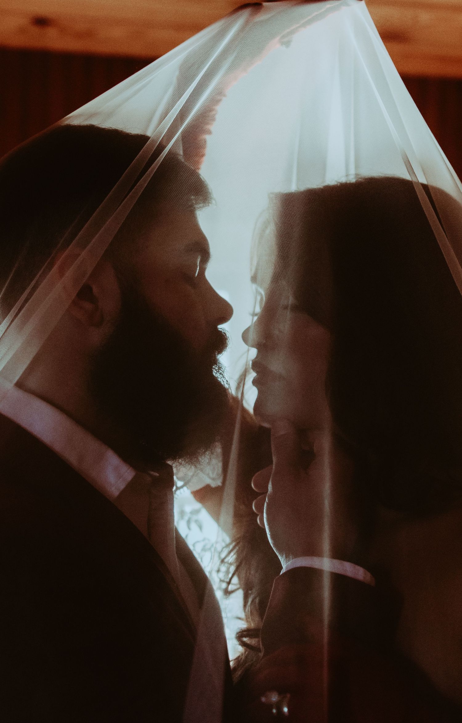 A bearded groom and bride share an intimate moment under a sheer wedding veil in warm, romantic lighting.