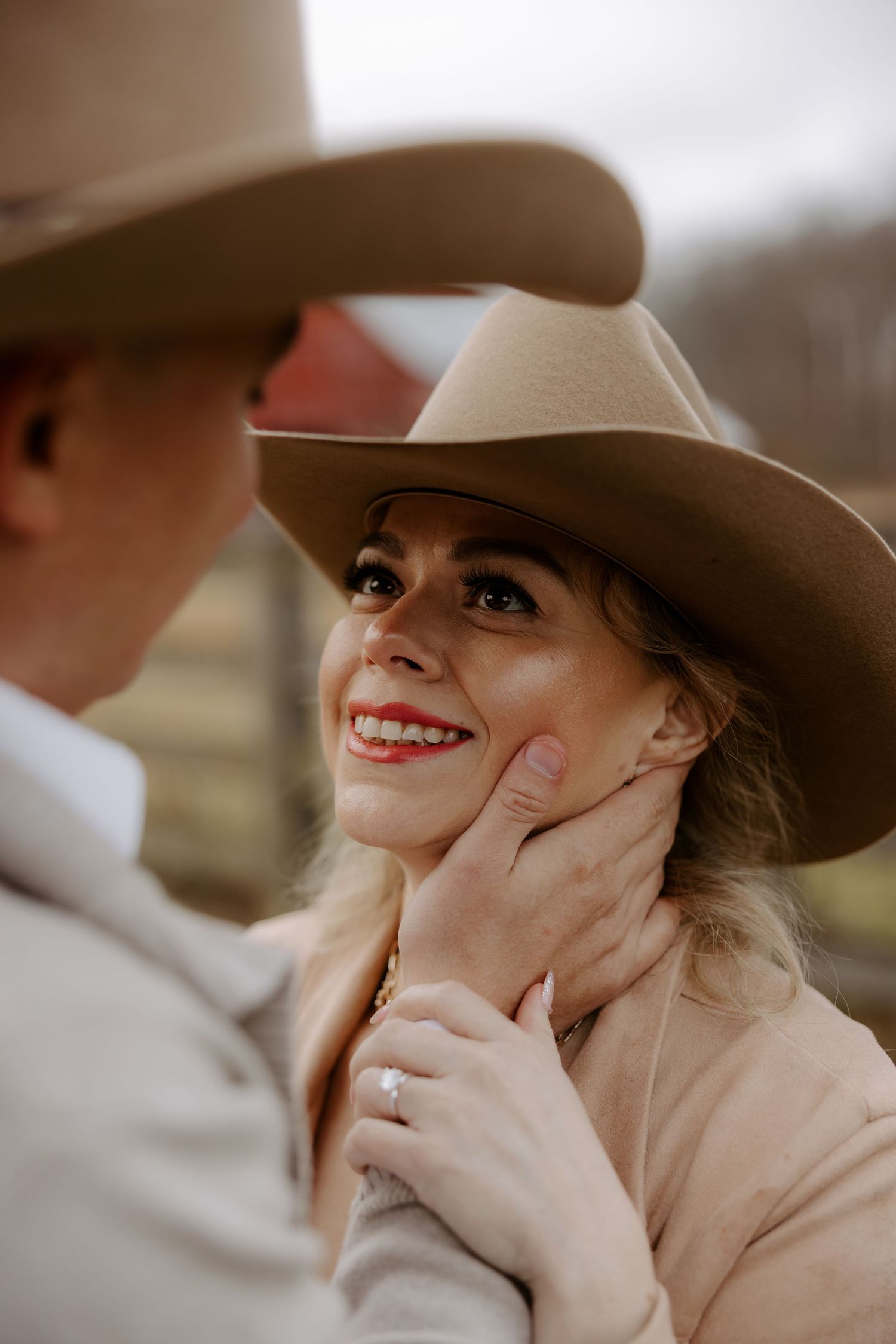 A smiling woman in a tan cowboy hat gazes up as a man gently cups her face, showcasing her diamond engagement ring.