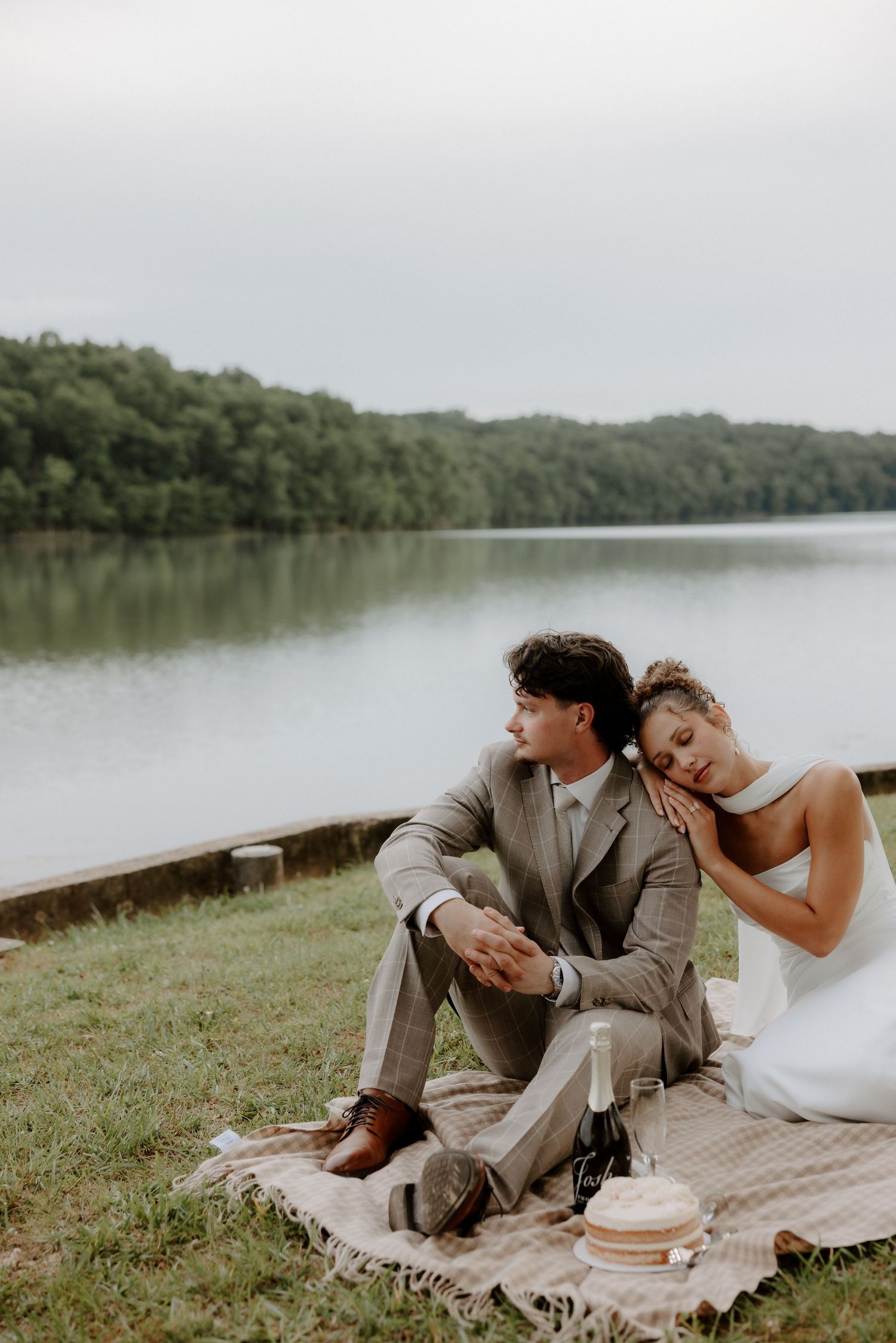 Couple relaxing on blanket by lakeside with champagne and cake during elopement.