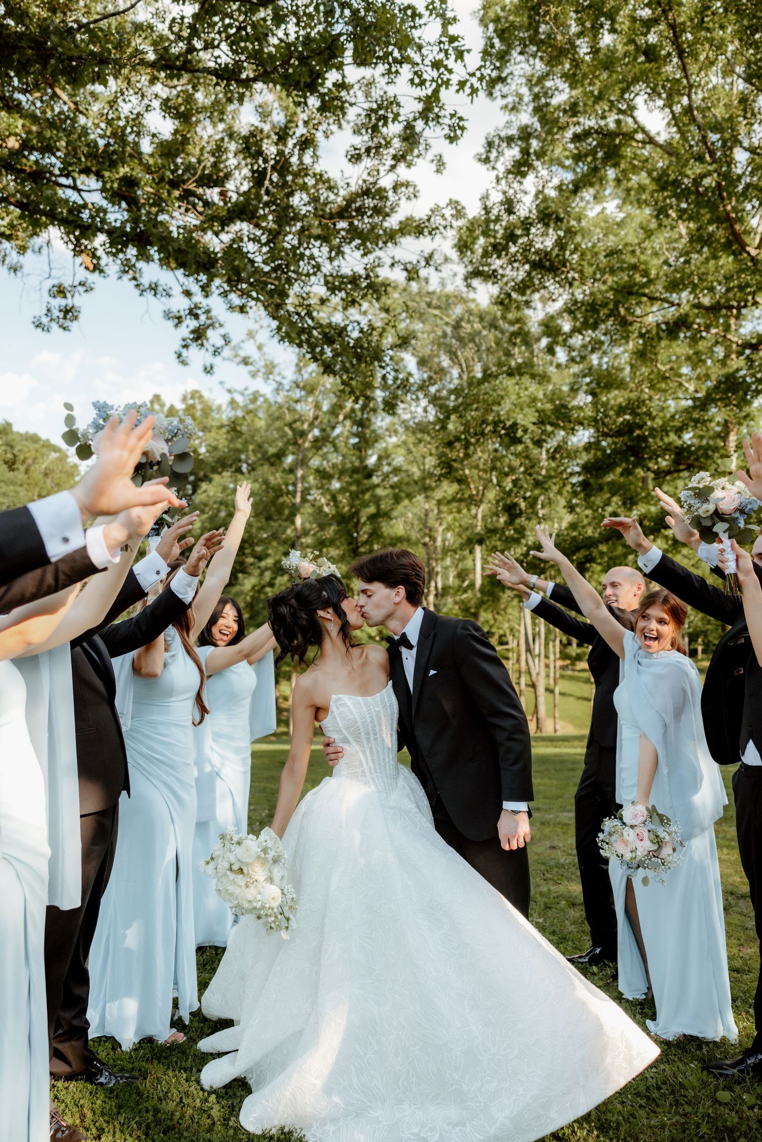 Bride and groom share a kiss under an arch of raised hands formed by their wedding party outdoors.