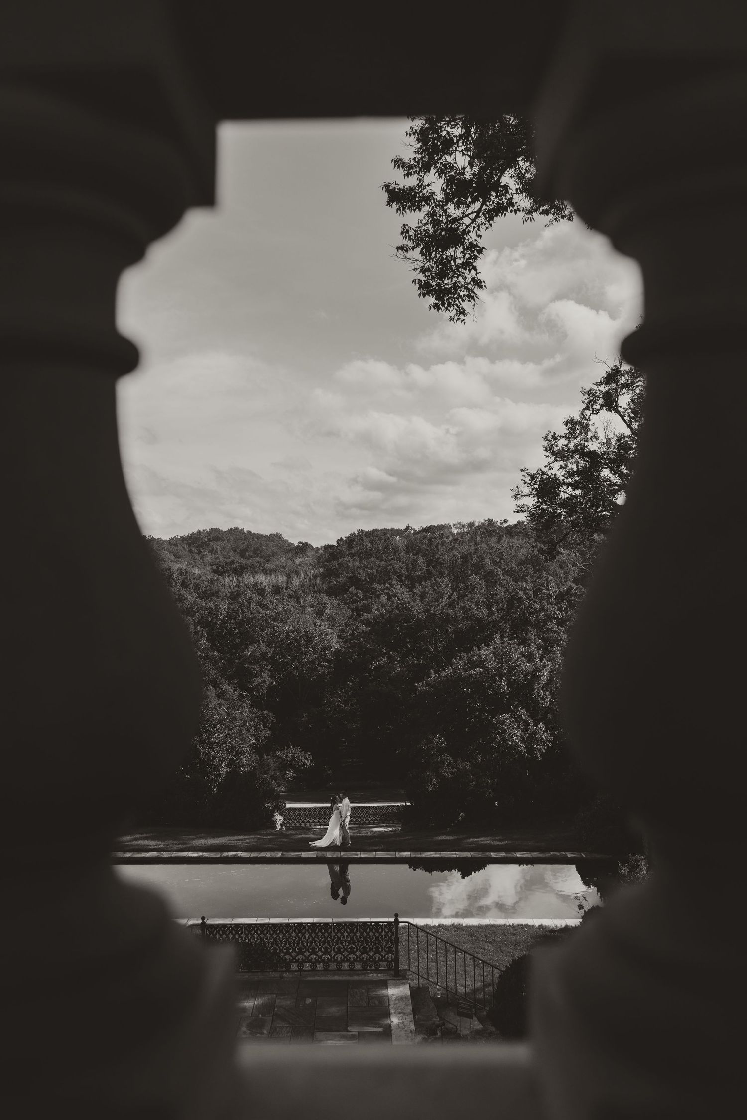Black and white photo of a bride framed through an ornate window opening, with a reflective pool and lush forested hills beyond.
