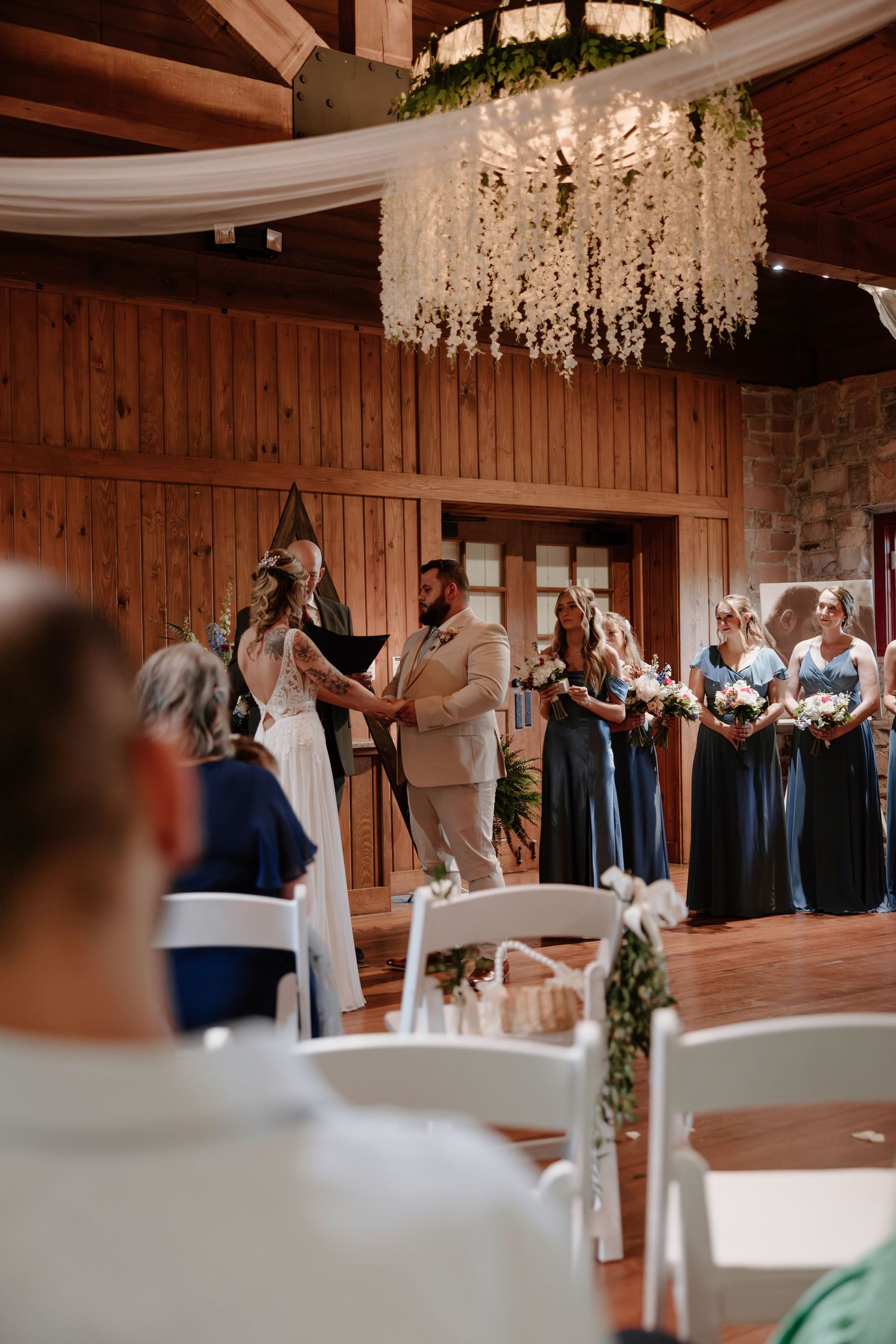 Wedding ceremony in progress under a floral chandelier in rustic wooden venue.