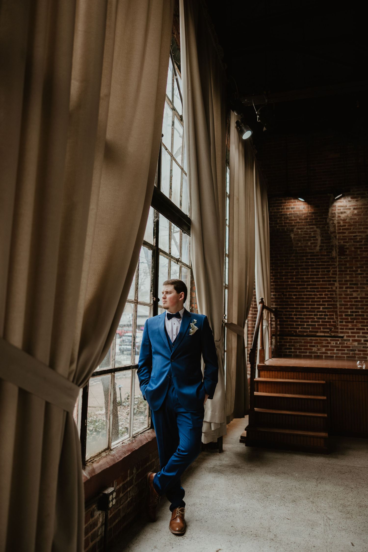 Groom in blue suit poses by tall industrial windows with dramatic lighting.