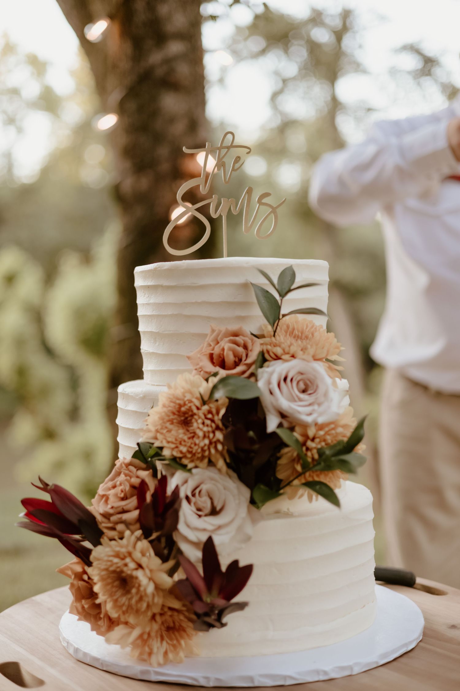 Elegant three-tier white wedding cake decorated with blush roses, burgundy flowers and peach dahlias in outdoor setting.
