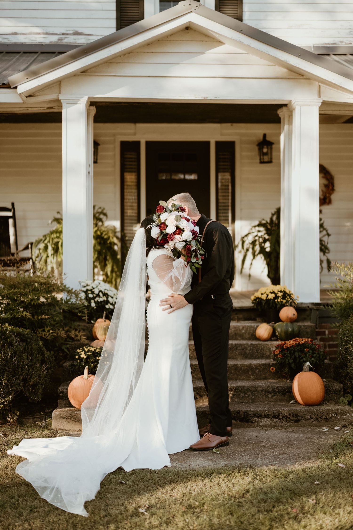 A couple shares an intimate moment on a white porch with fall pumpkin decor and floral bouquet at their wedding.