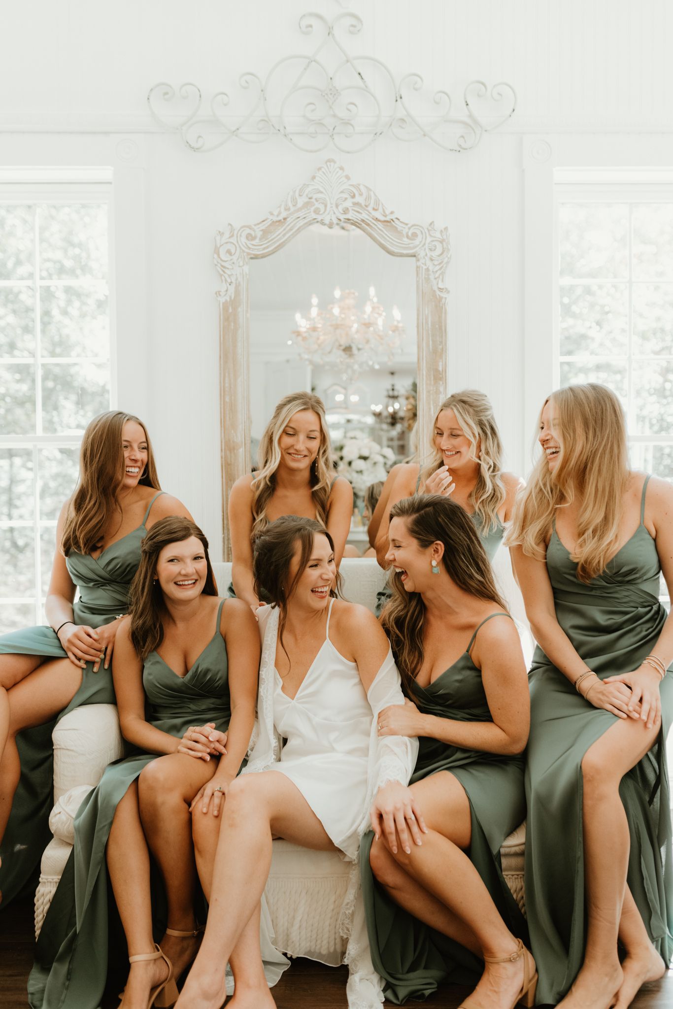 Wedding party shares candid laughs while sitting together on an elegant white couch in a bright sunlit room.