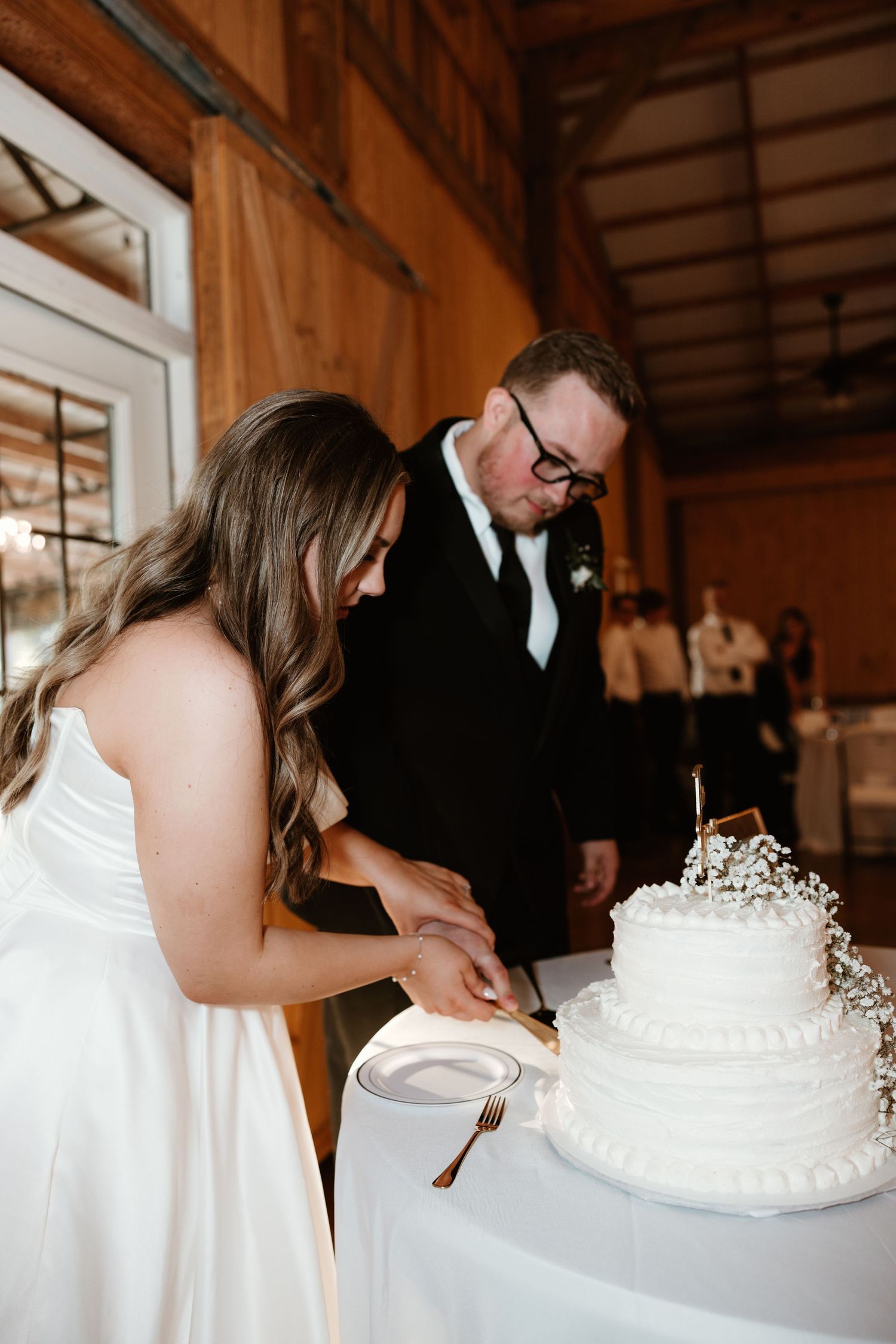 Couple cuts into their elegant white wedding cake with delicate pearl details at rustic wooden reception venue.