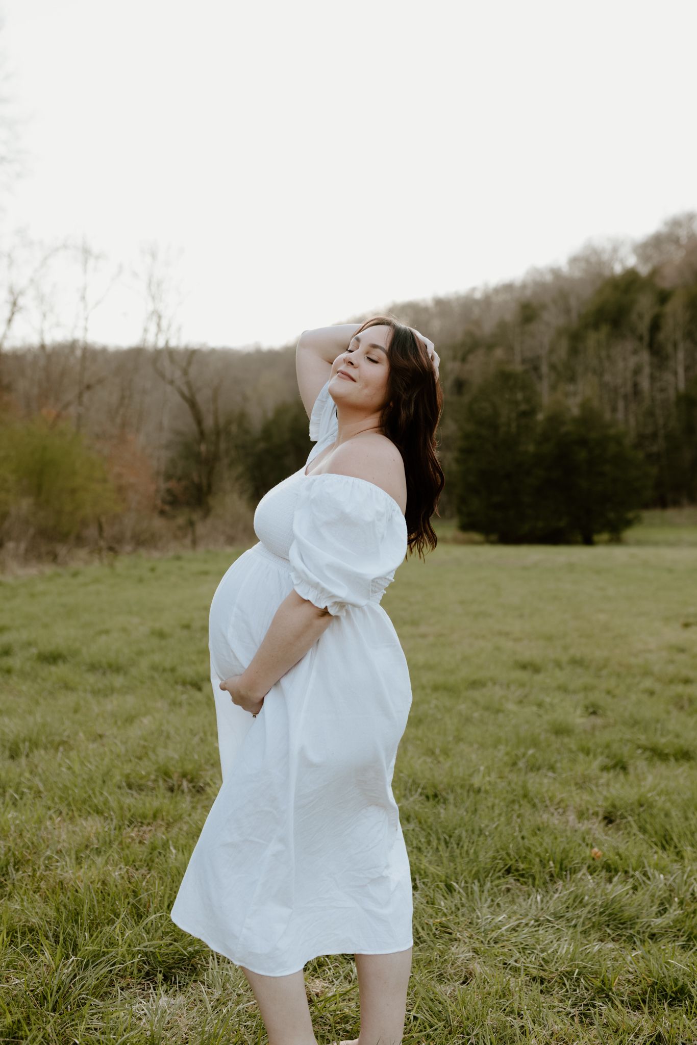 Pregnant woman in white off-shoulder dress smiles while posing in a scenic outdoor field setting.