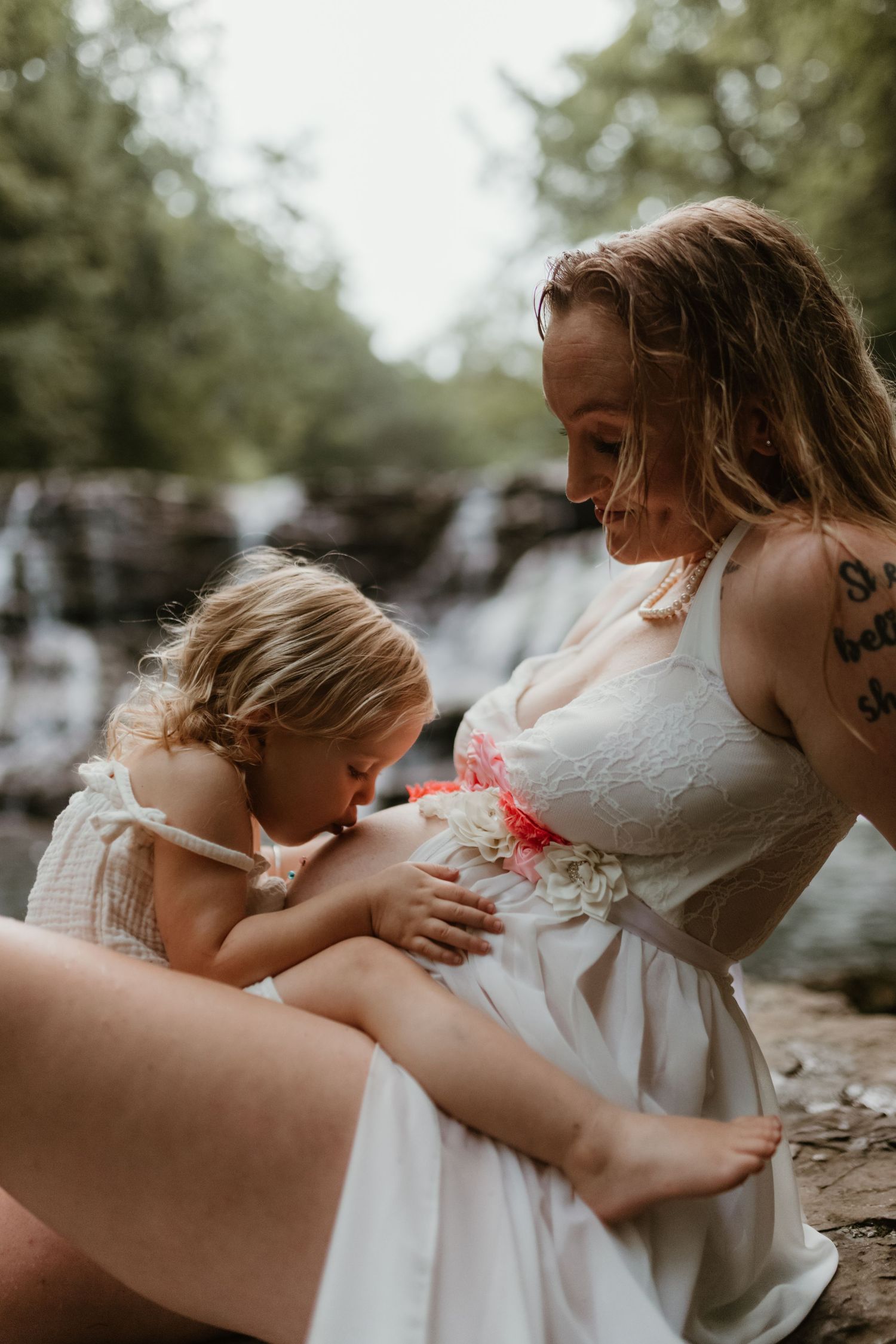 An intimate maternity moment captured by a waterfall as sunlight filters through the trees.