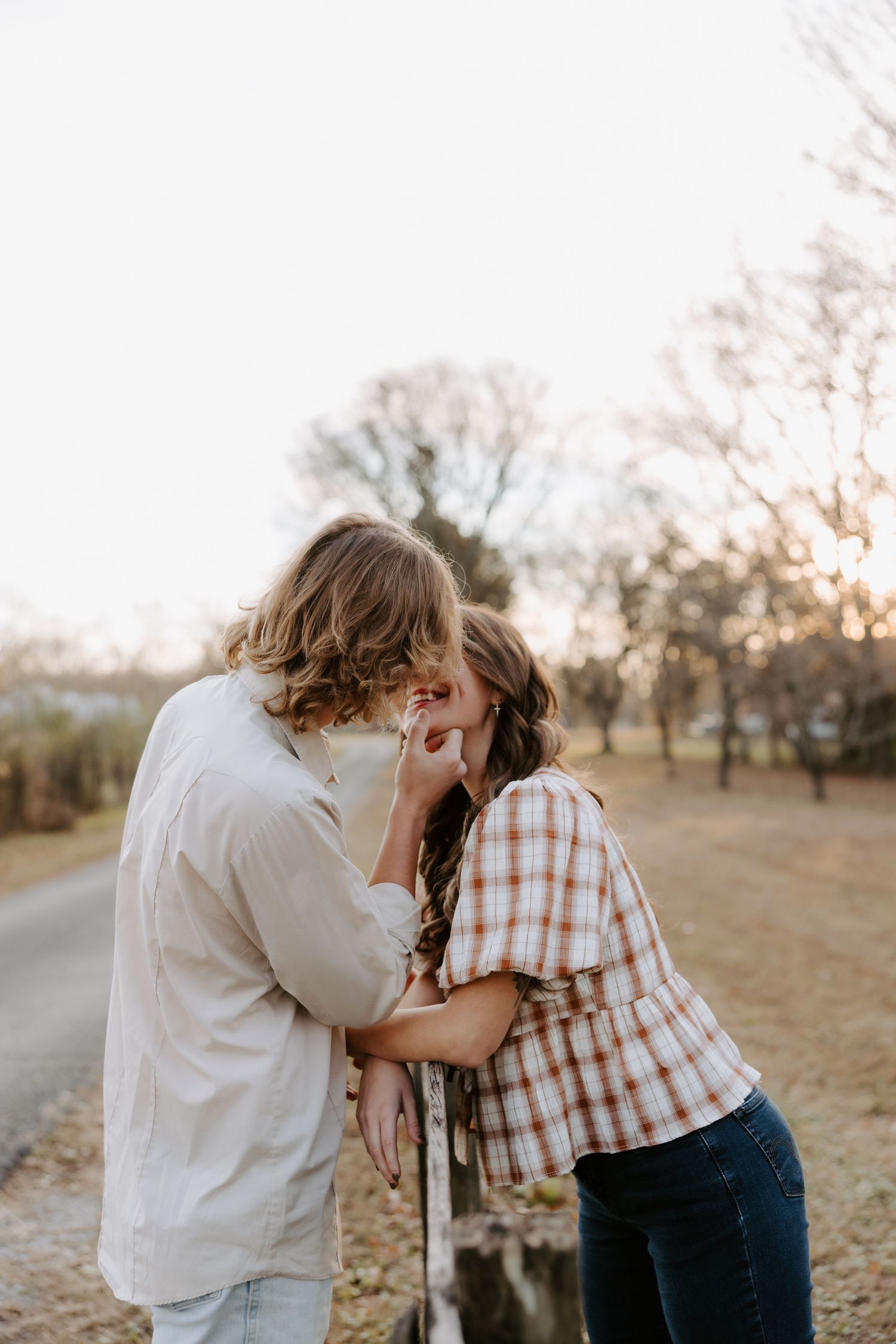 A couple shares a romantic moment leaning on a fence at golden hour in a rustic outdoor setting.