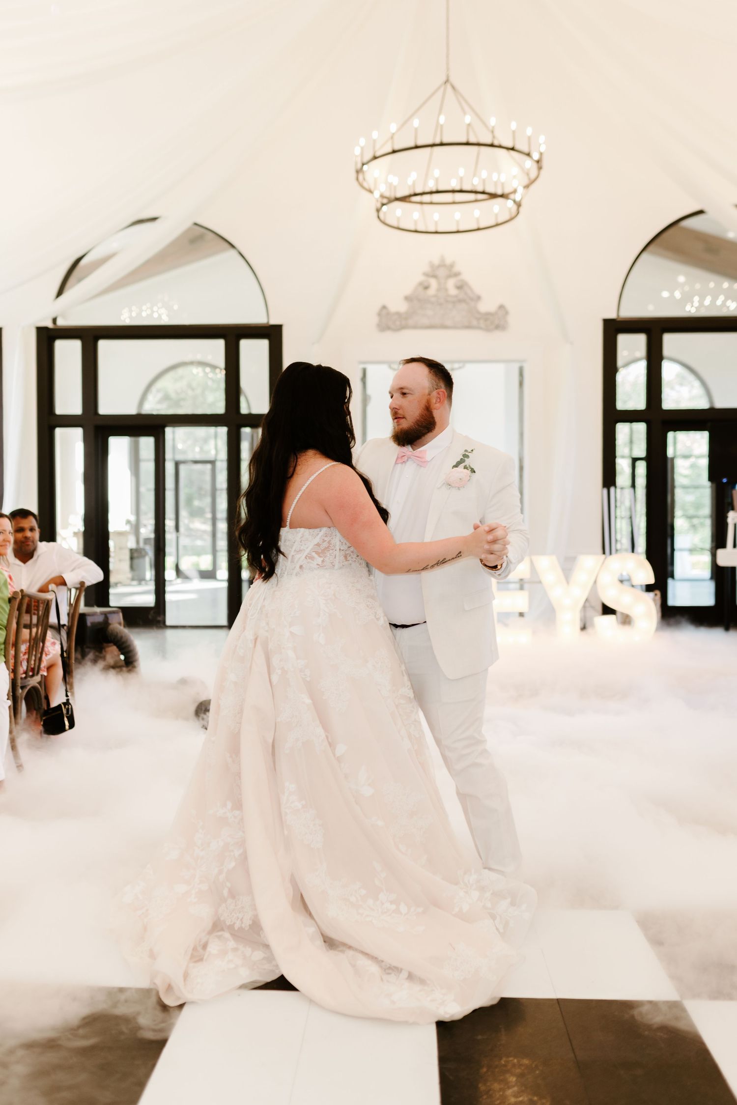 Bride and groom share their first dance on a foggy white dance floor beneath a chandelier in an elegant white venue.