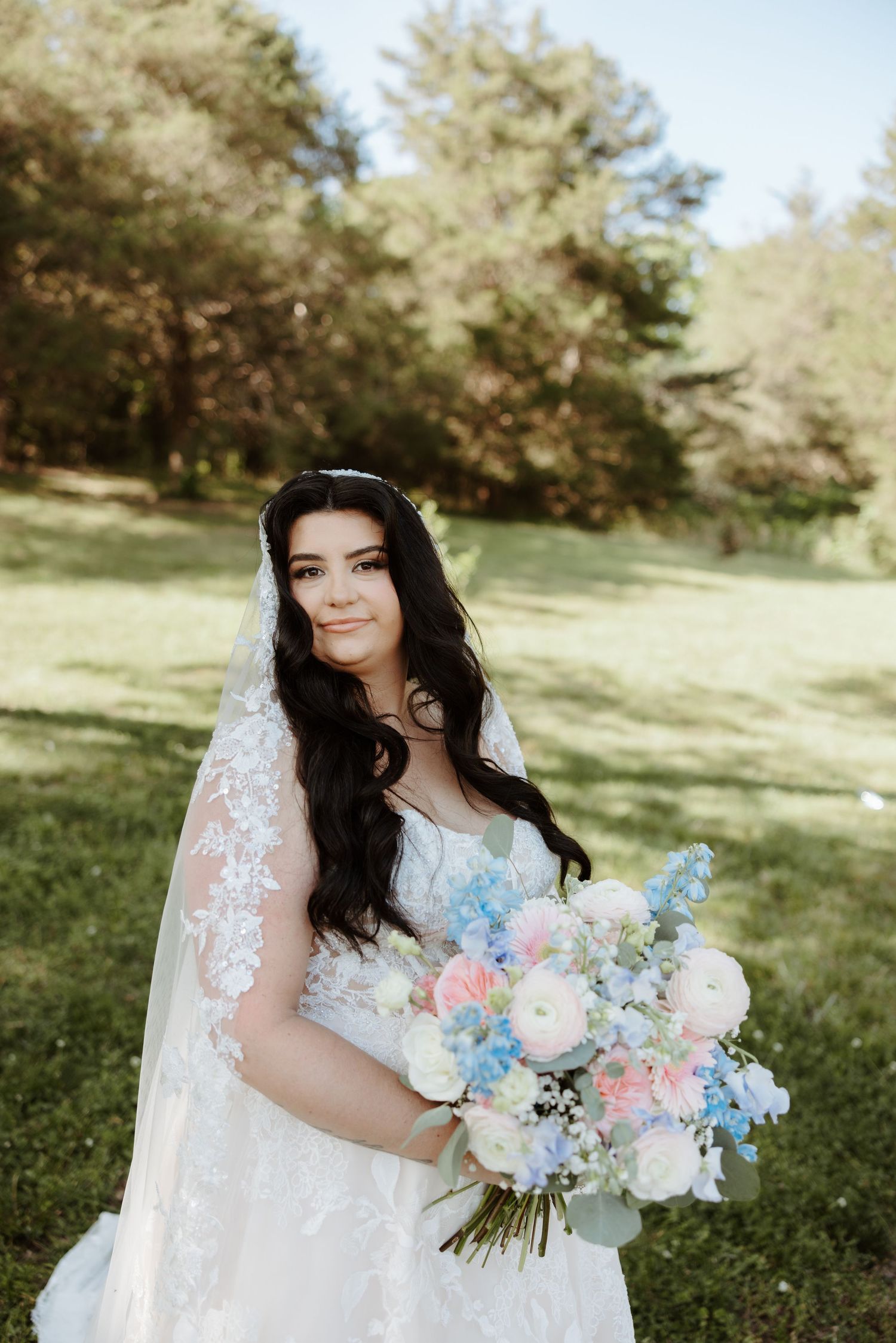 Bride in lace wedding gown and veil holds pastel bouquet of pink, white, and blue flowers outdoors.