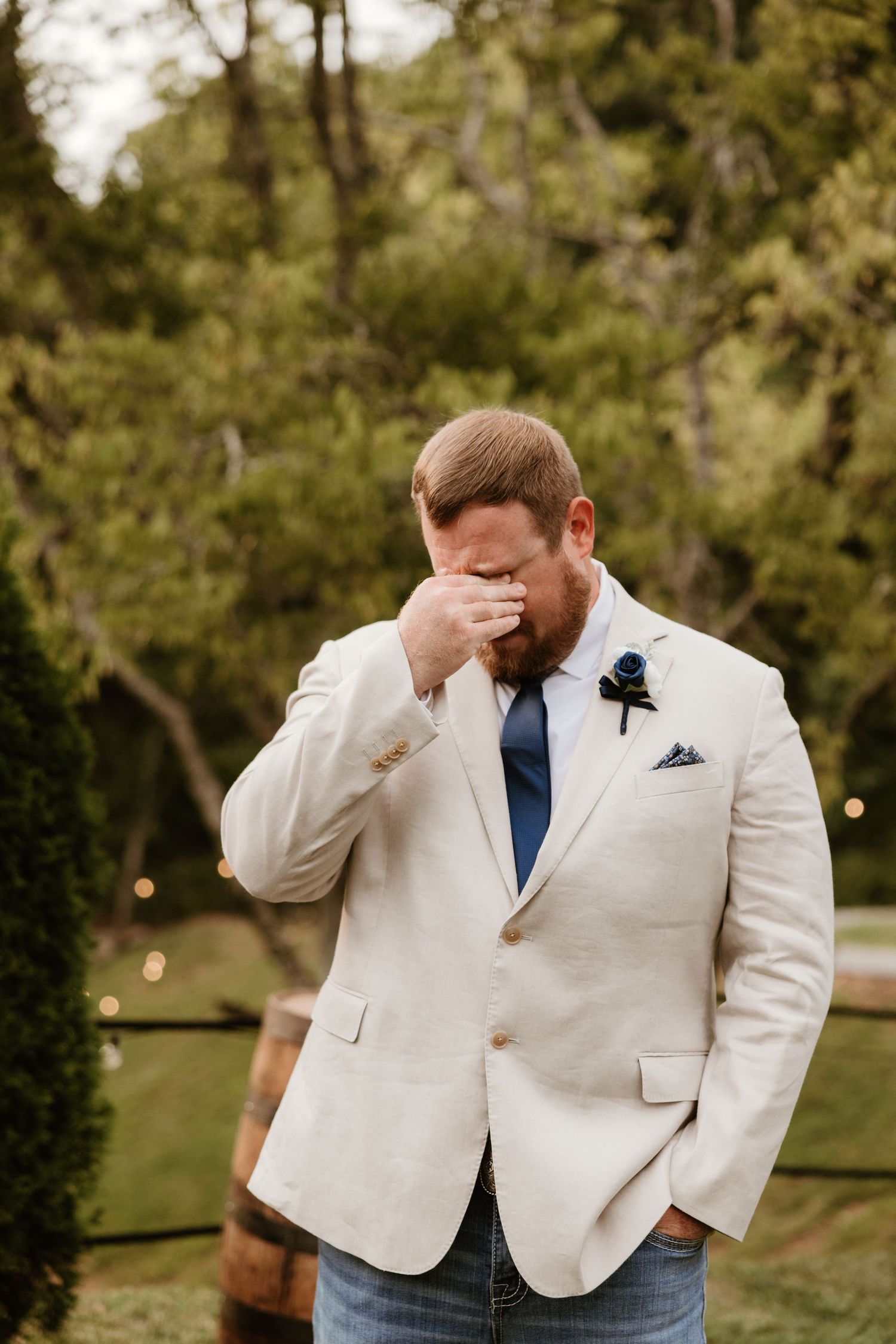 A person in a white suit jacket and navy tie reacts emotionally during a wedding ceremony in an outdoor garden setting.