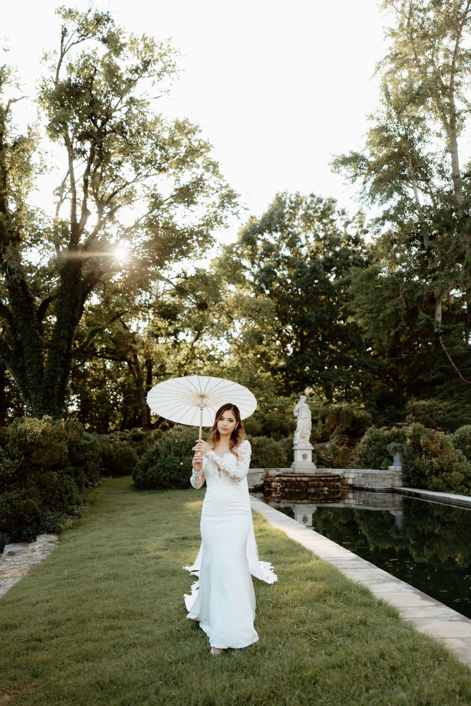 Bride in white gown holding parasol umbrella in lush garden with reflecting pool and statue at golden hour.