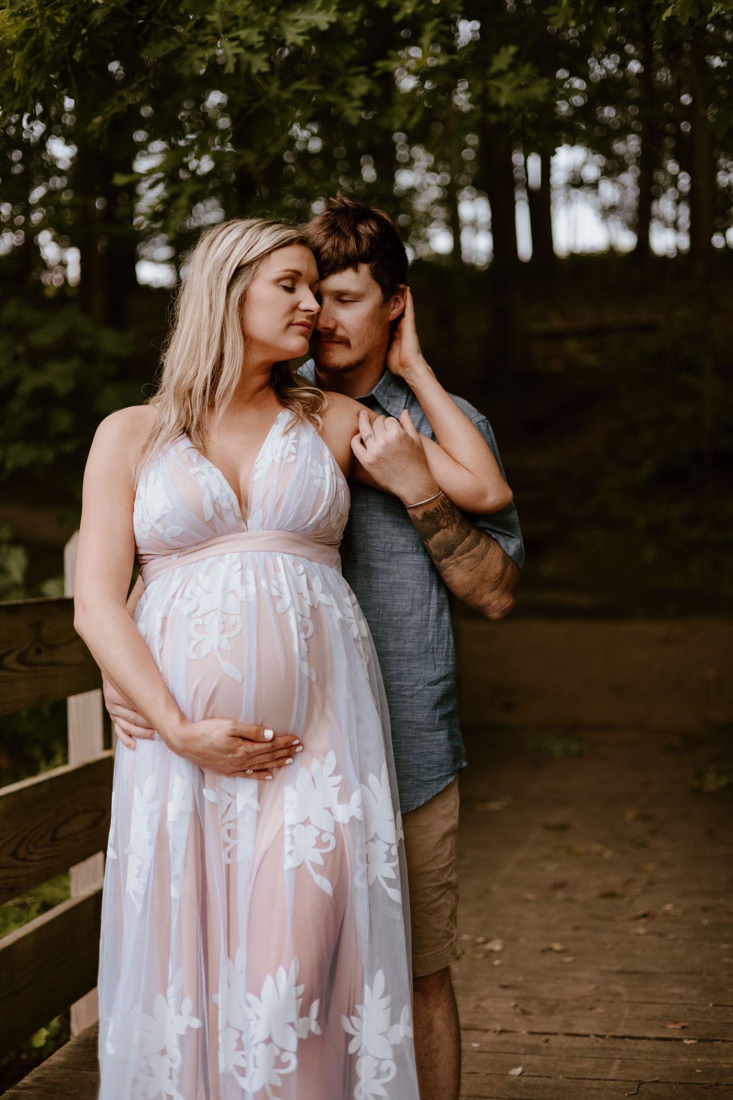 An expecting couple shares an intimate moment outdoors in a wooded setting wearing white lace and casual attire.