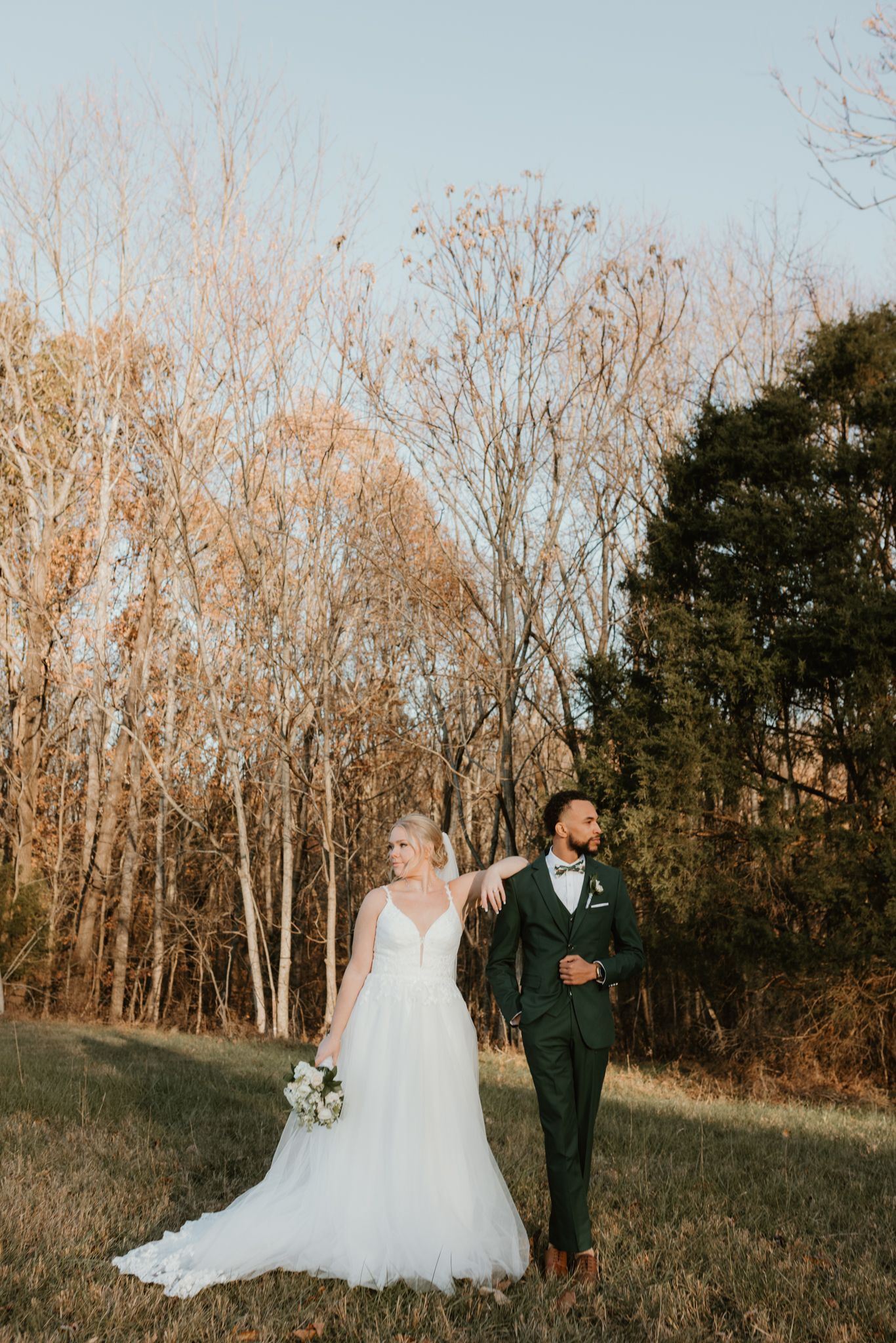 A wedding couple poses elegantly against autumn trees in a rustic outdoor setting.