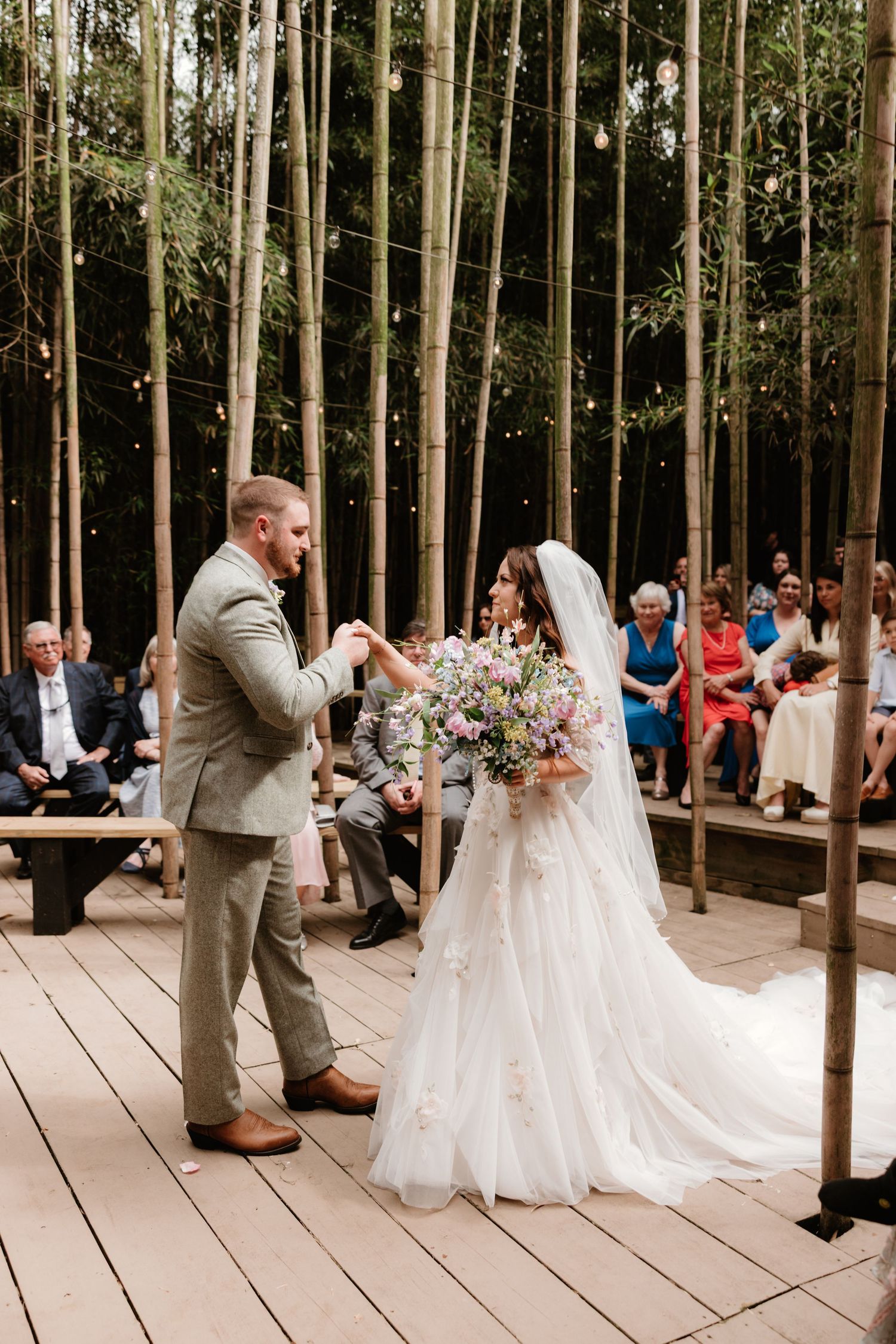 A wedding ceremony takes place on a wooden deck surrounded by tall trees and wedding guests.