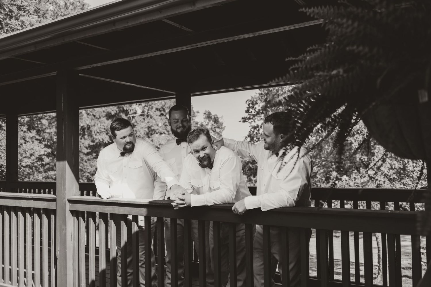 A vintage black and white photograph shows three people leaning against a wooden porch railing outdoors.