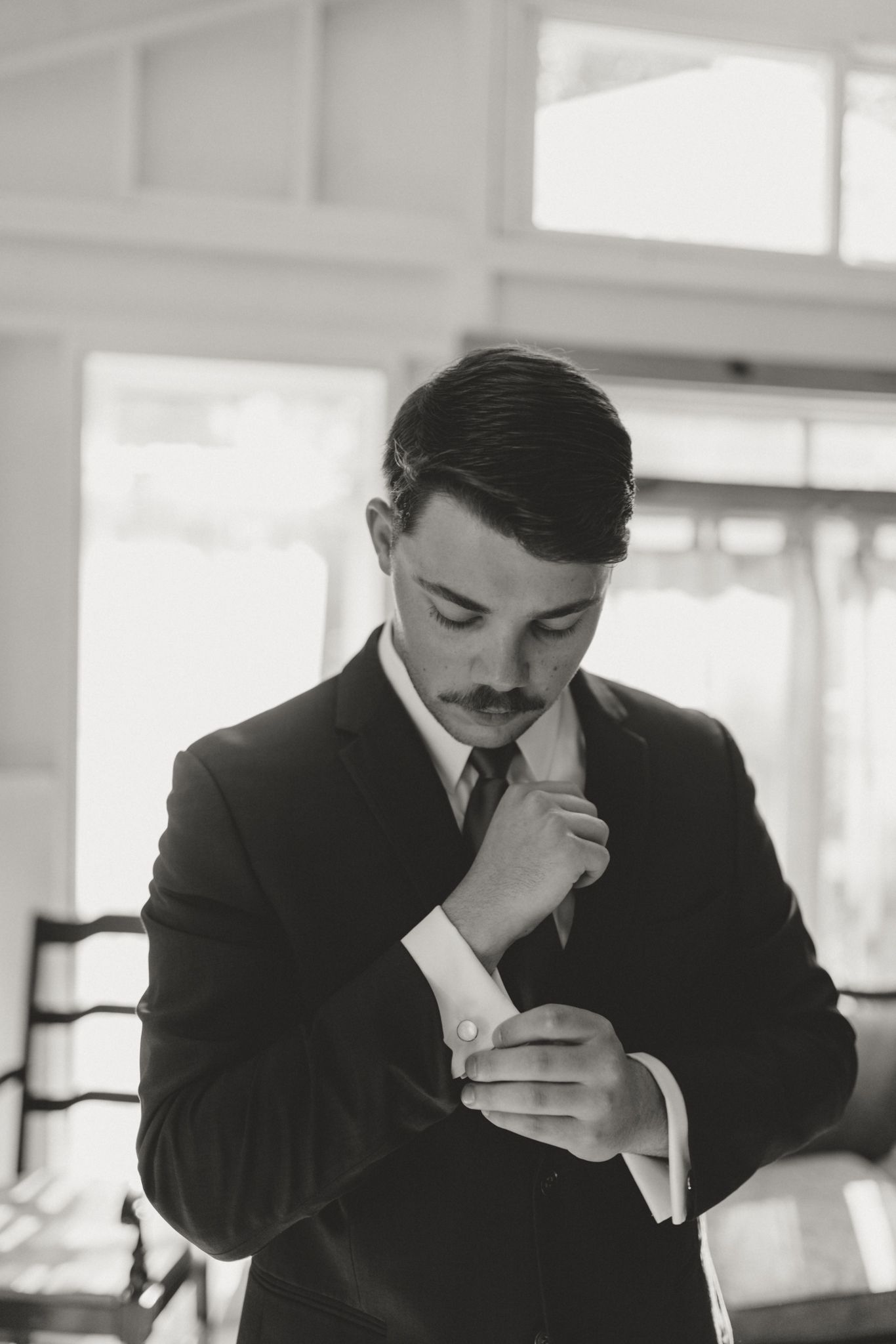 Classic black and white portrait showing figure in suit adjusting cufflinks in elegant interior setting.