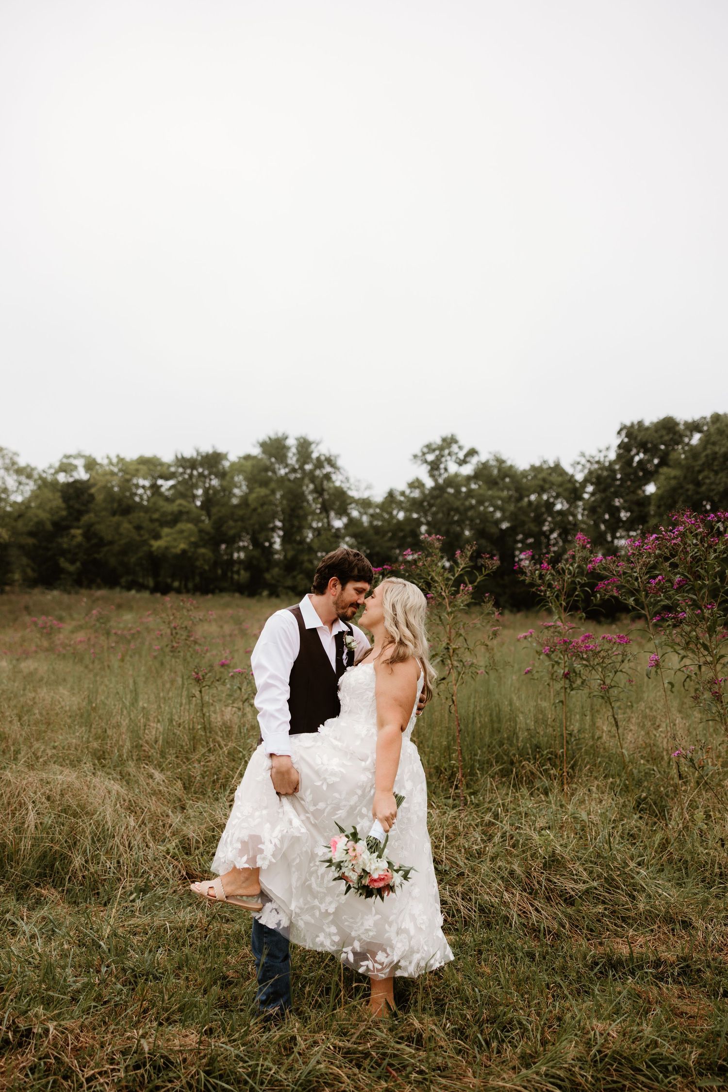 A couple in wedding attire stands in a rustic field with blooming wildflowers during a summer sunset photo session.