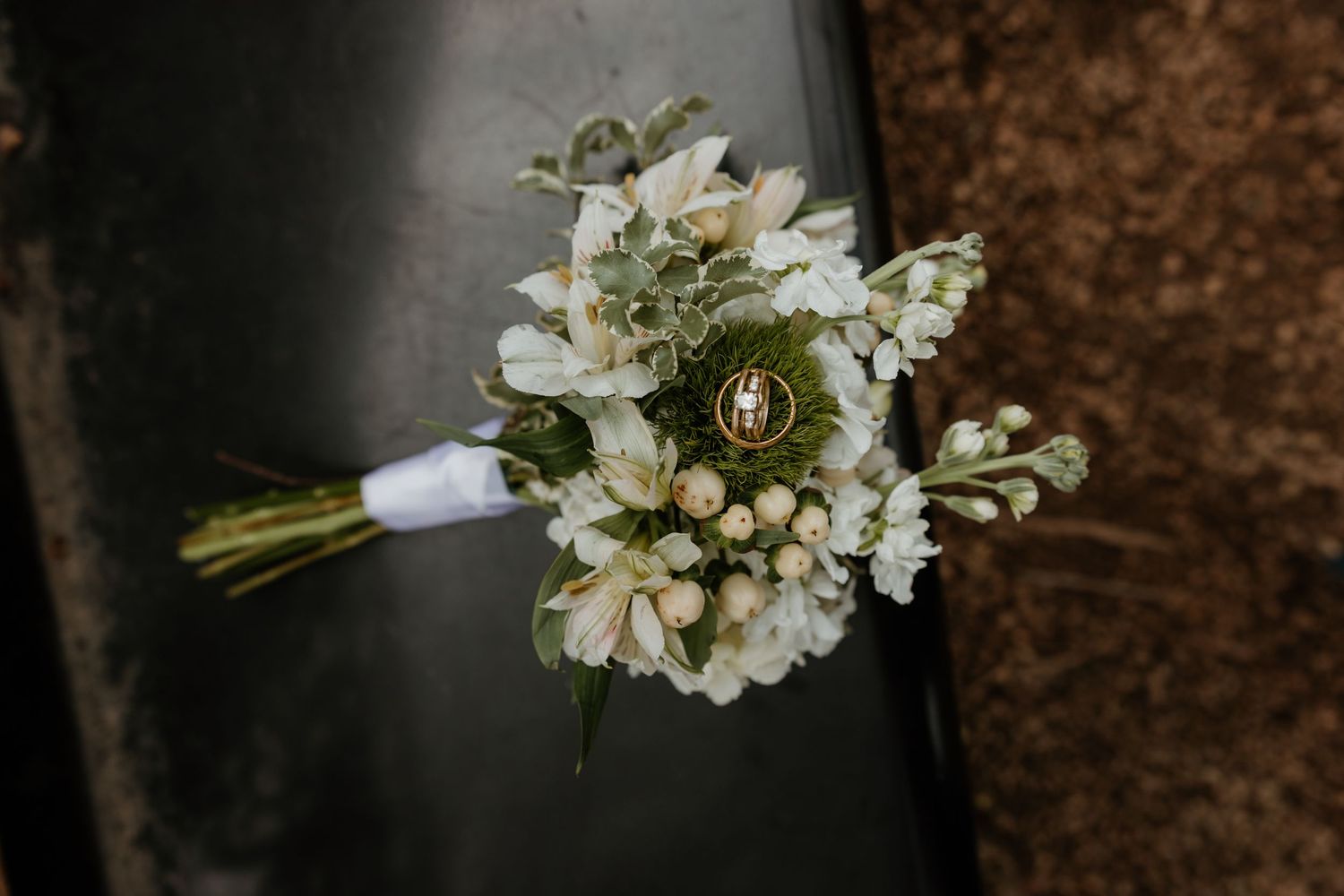 White wedding bouquet with delicate blooms and rings nestled in greenery, resting on a dark surface.