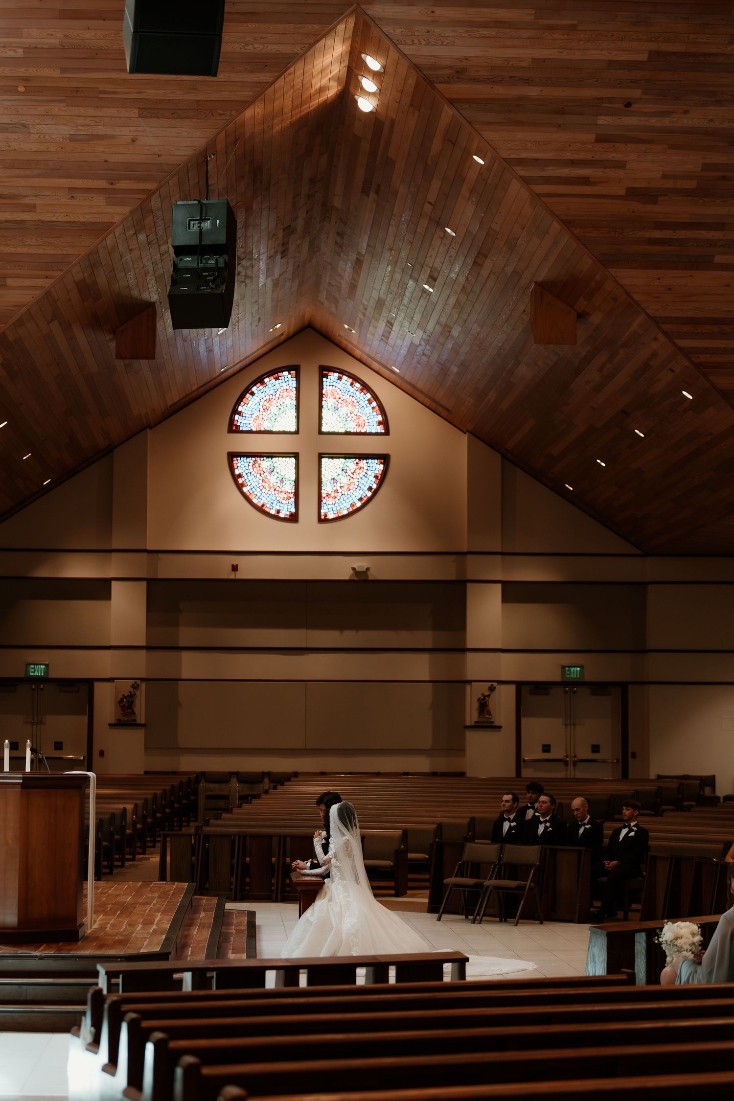 Bride walks down the aisle in a church with wooden ceiling and stained glass cross window during wedding ceremony.