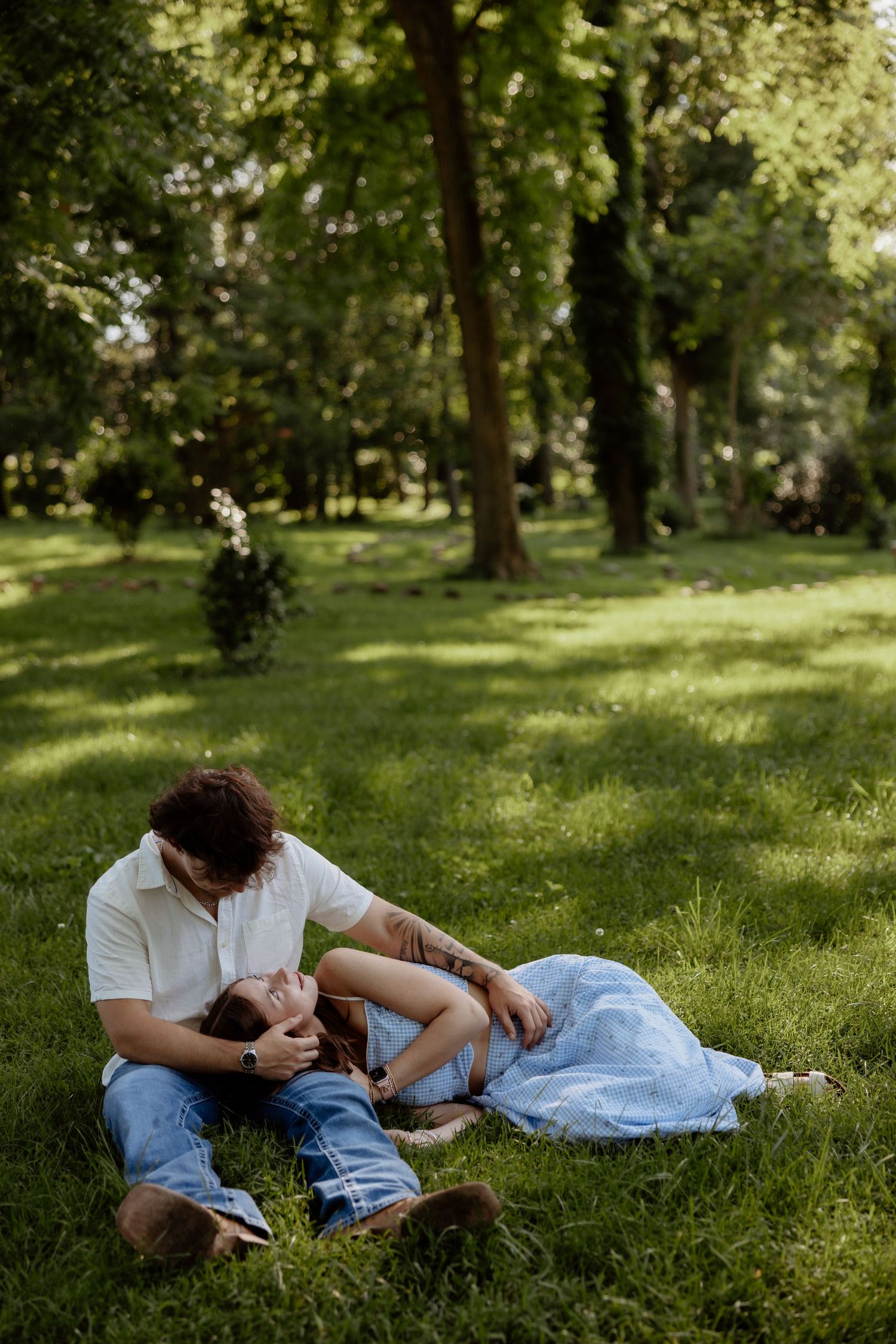 Romantic couple relaxing on green grass in a sunny park, woman lying in man's lap wearing a blue dress.