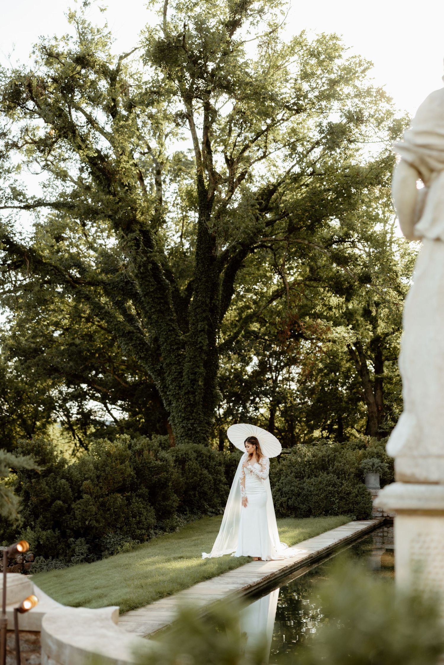 Bride in white gown and sun hat stands by a reflecting pool in a lush garden with large trees.