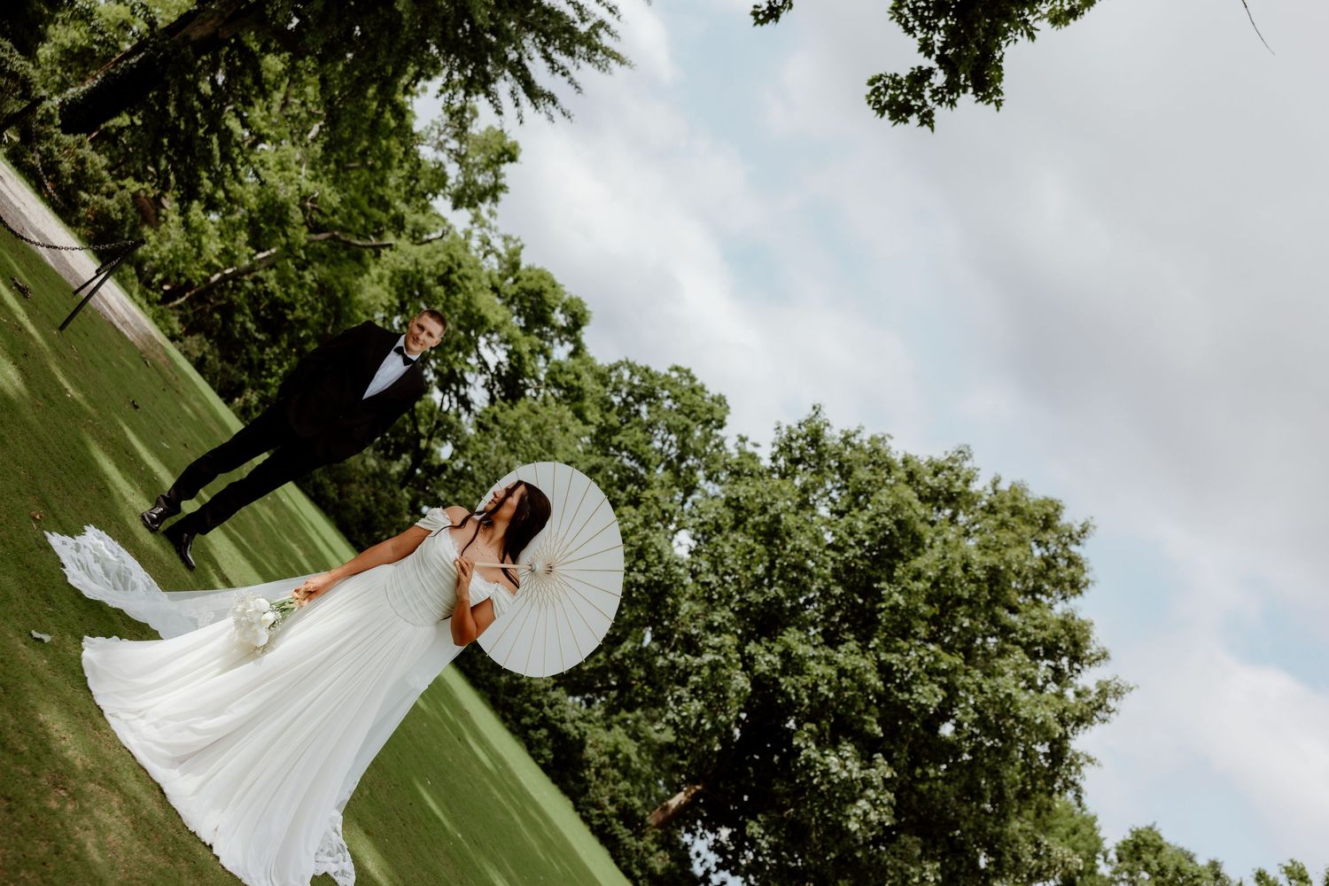 Bride in white gown holding parasol stands with groom in black suit on lush green lawn at outdoor wedding.