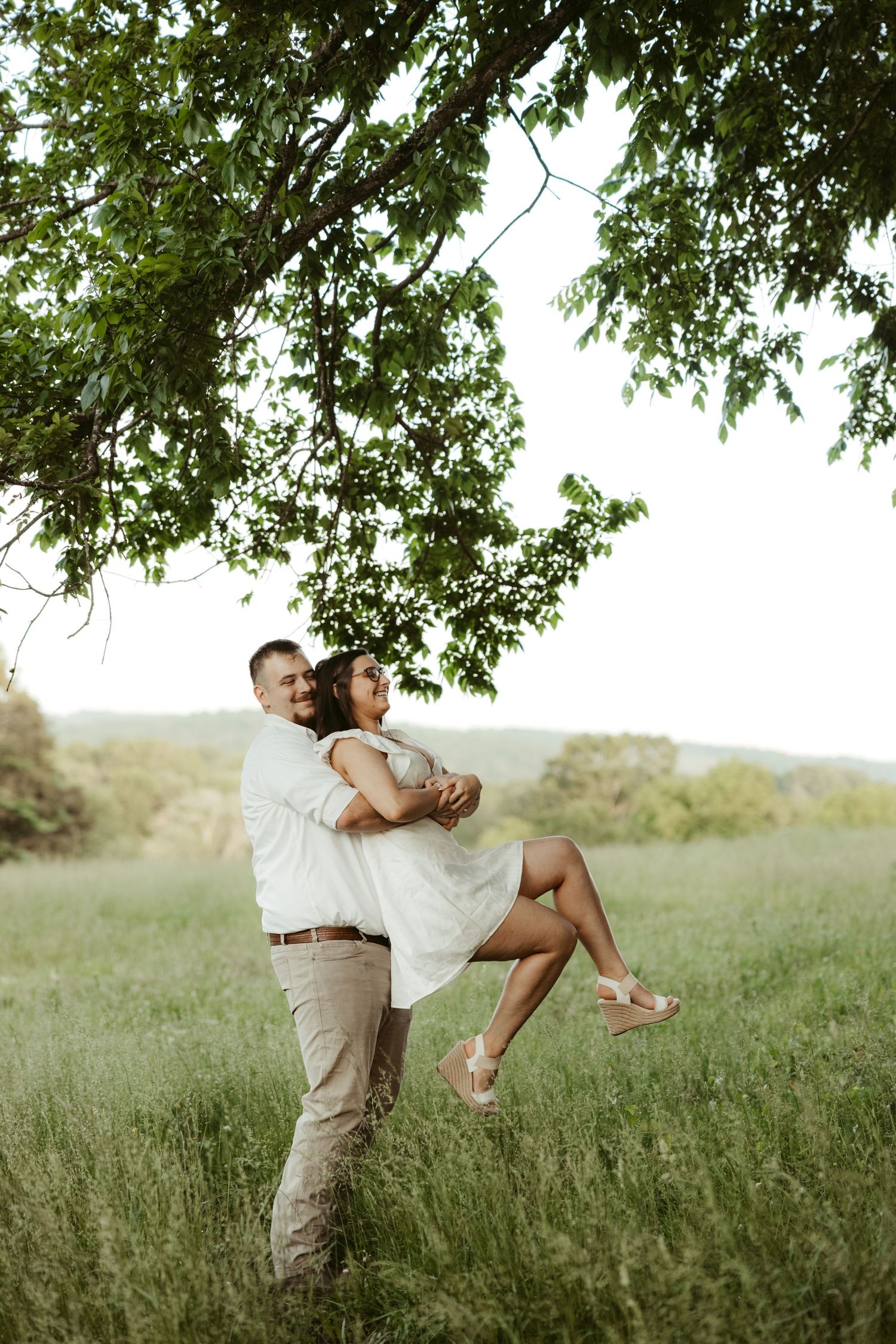 Couple embracing in golden field under tree branches during sunset photoshoot.