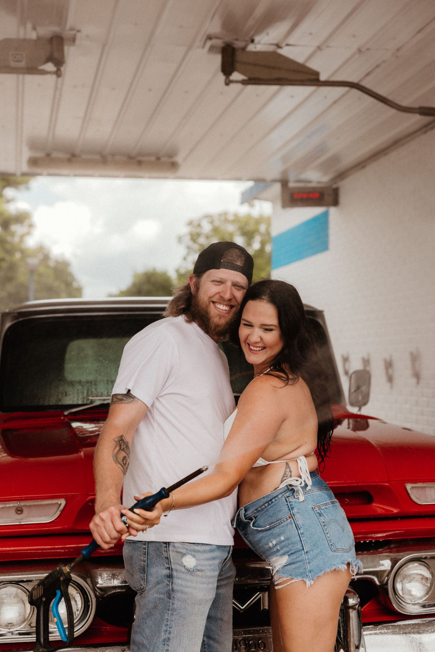 A joyful couple poses in front of a vintage red GMC truck at a car wash.