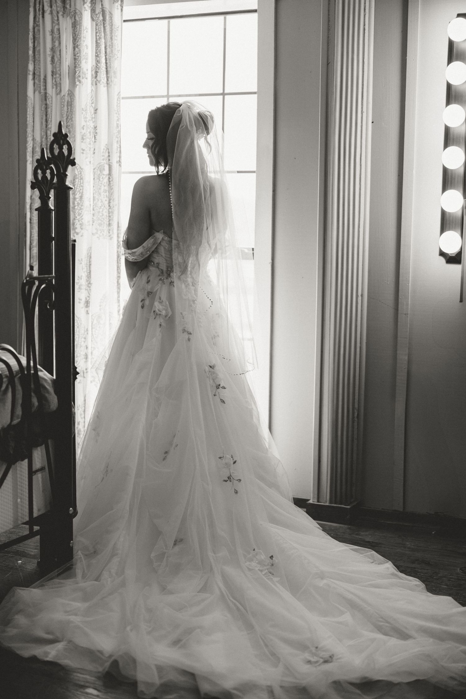A bride in a flowing white wedding gown with delicate embellishments stands silhouetted against a bright window.