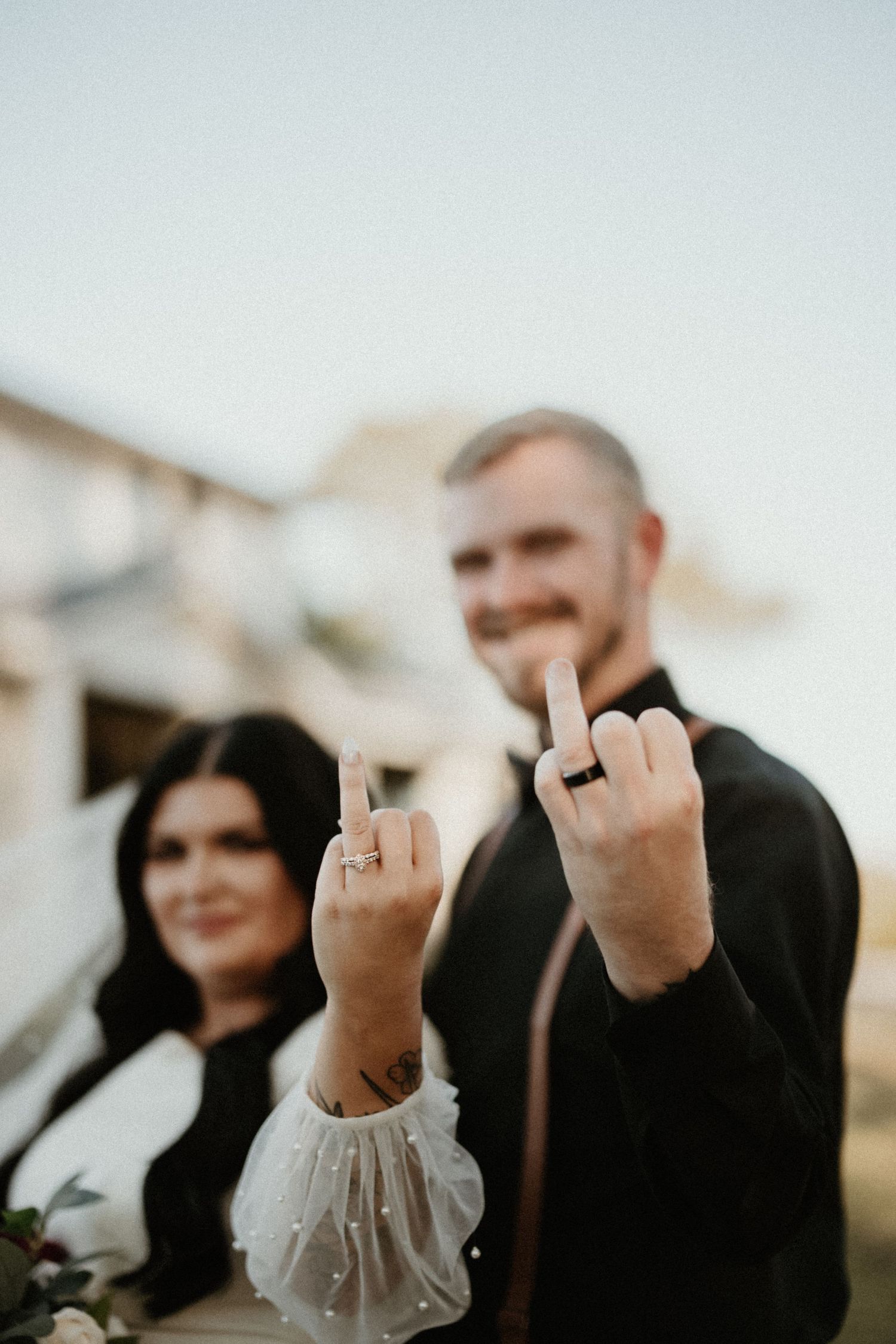 A playful wedding couple showing their middle fingers to the camera while smiling outdoors.