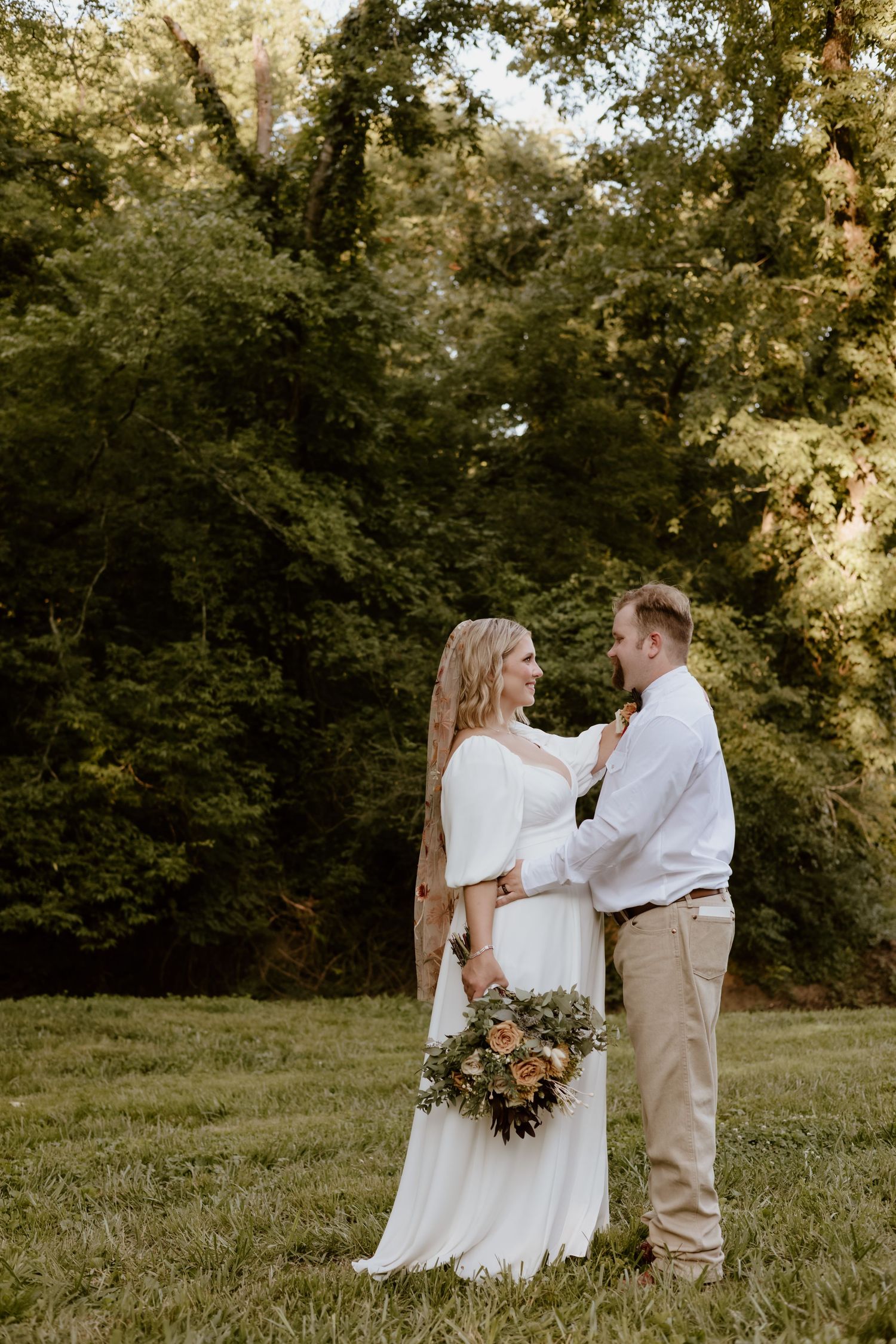 Rustic outdoor wedding portrait with a couple in casual attire standing together on a grassy hillside.