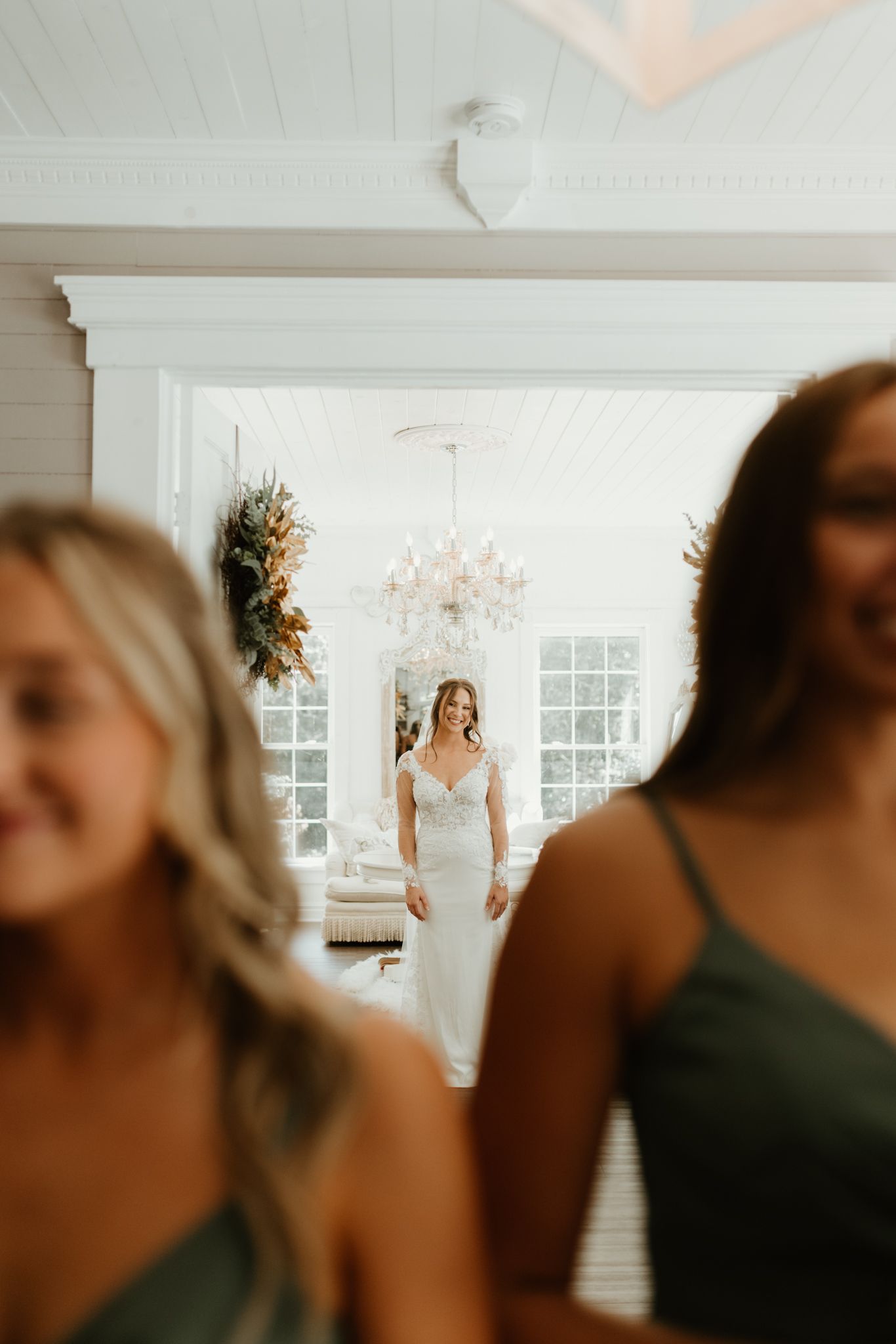 A bride in a white lace gown stands elegantly in a bright room with a crystal chandelier while guests gather in front.