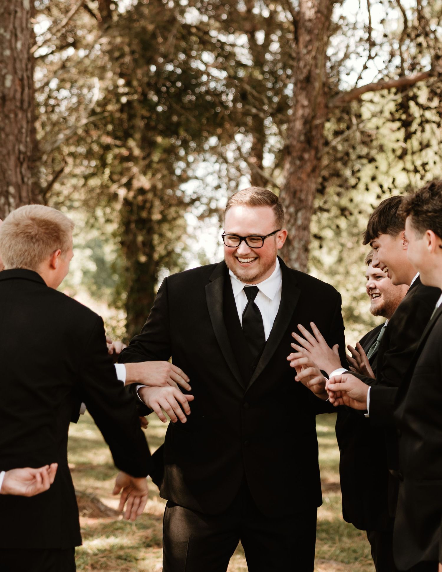 Wedding party members in black suits share laughter and playful moments during outdoor photo session.