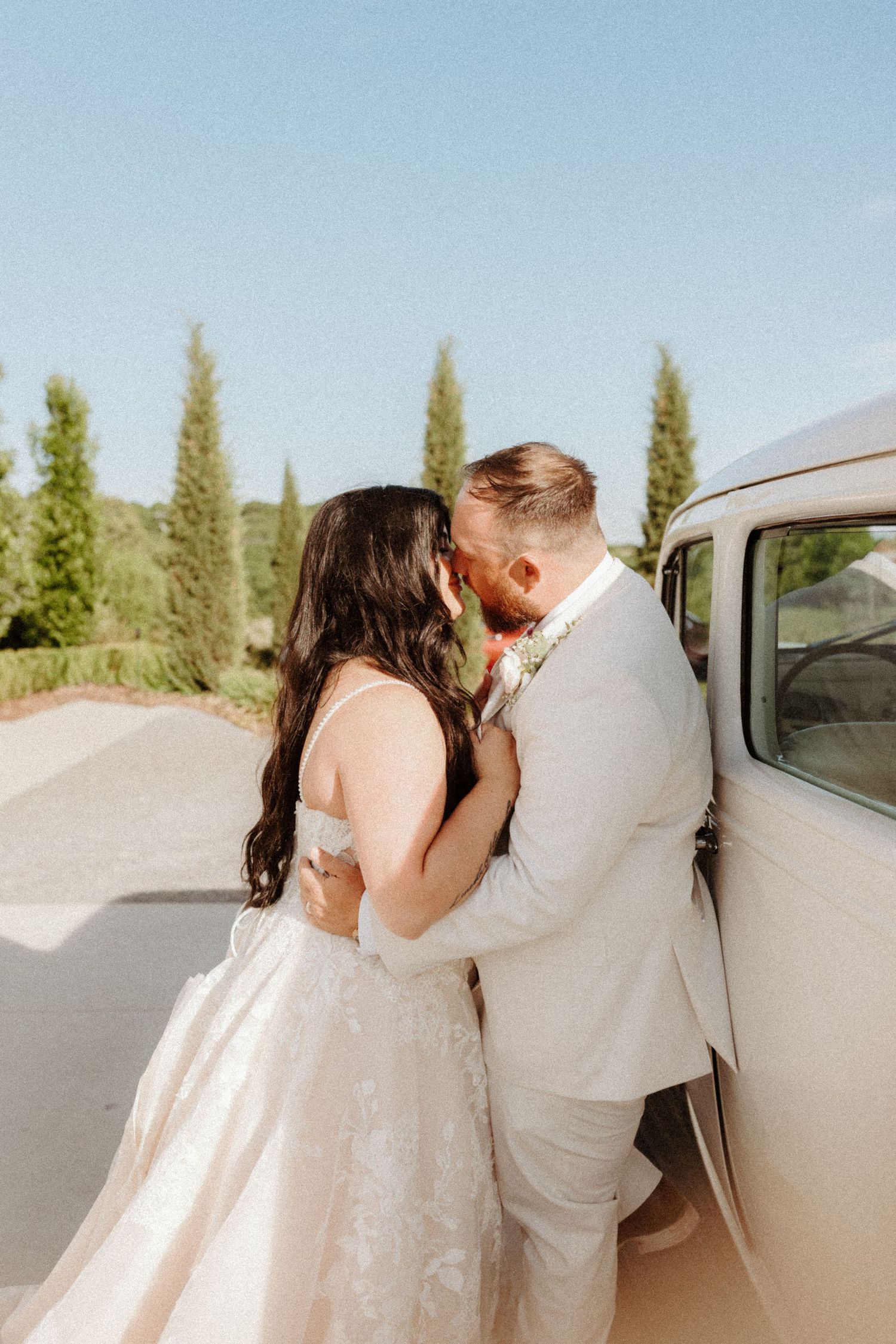 Bride and groom share a kiss leaning against a vintage white car with cypress trees behind them.