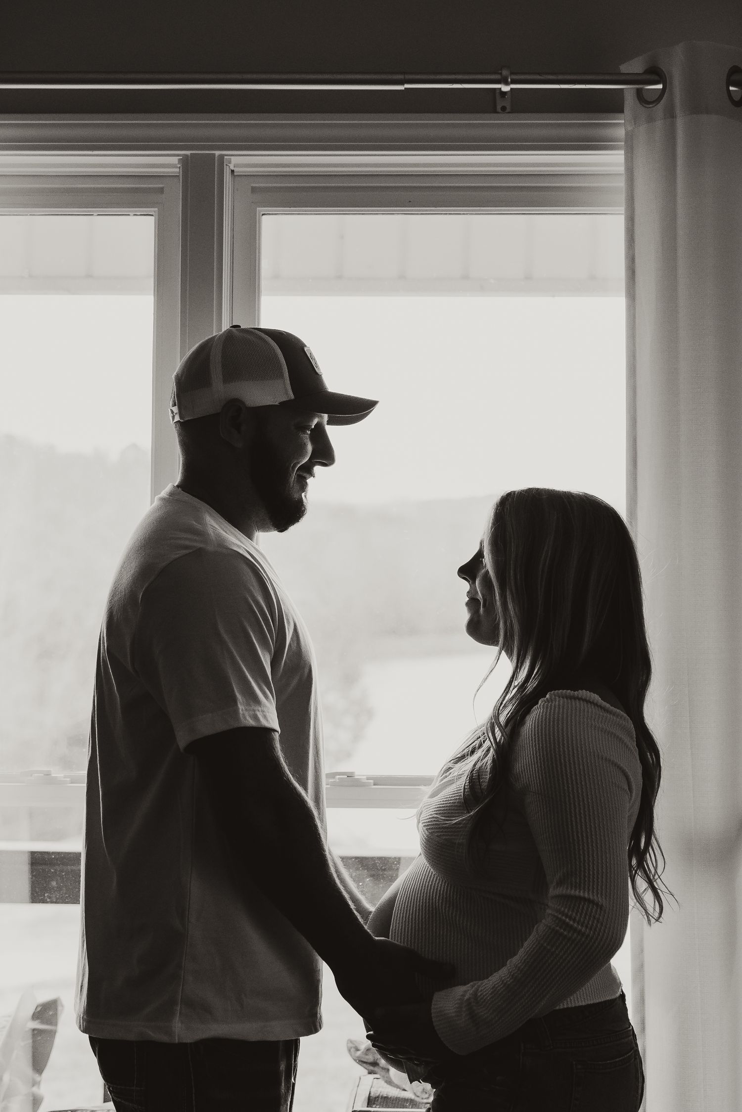 Black and white photo of a pregnant couple holding hands, silhouetted against a bright window in a tender moment.