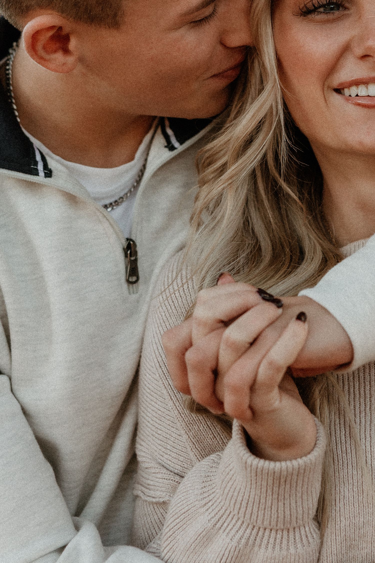 A couple holds hands tenderly while sharing a kiss, dressed in cozy neutral sweaters in a romantic close-up photo.