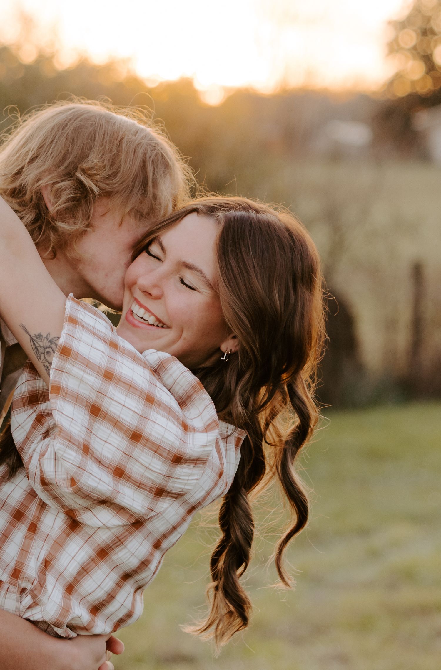 A joyful couple shares a tender embrace outdoors at golden hour, with the woman smiling brightly as her partner hugs her from behind.
