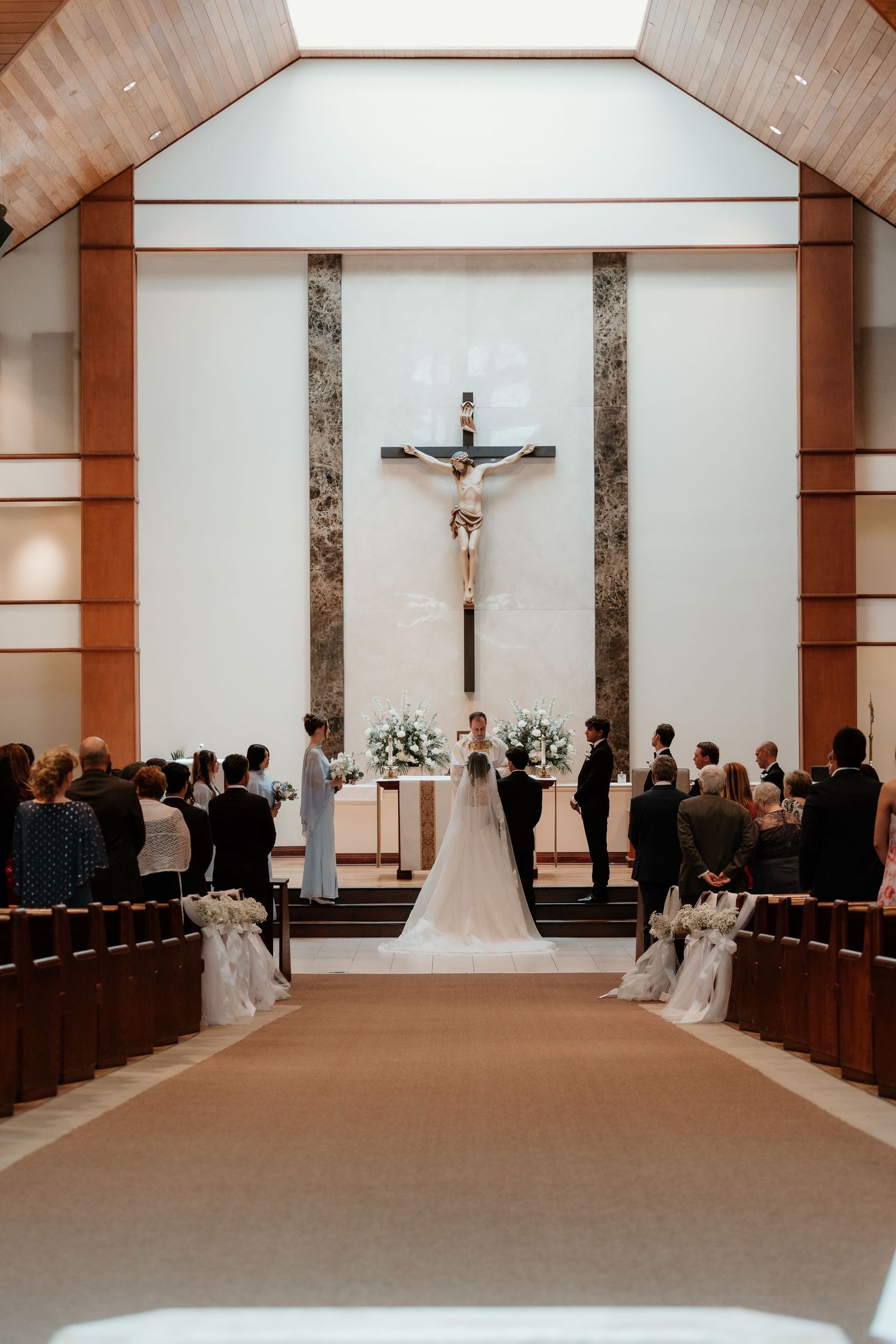 A bride and groom exchange vows at a Catholic church altar beneath a crucifix, surrounded by guests seated in wooden pews.